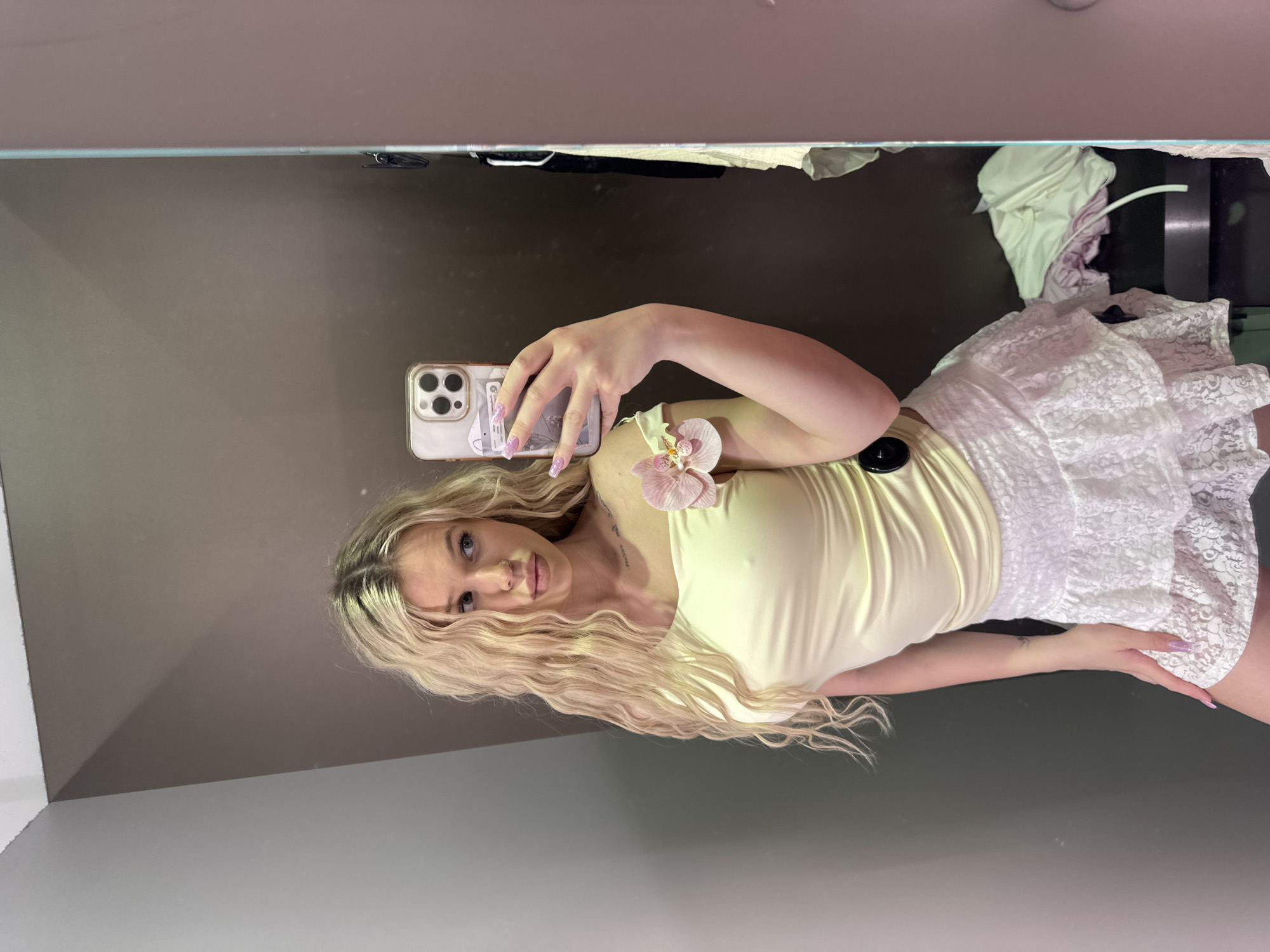 A young woman with long, wavy blonde hair is taking a selfie in a fitting room. She is wearing a light yellow off-the-shoulder top and a white lace skirt. A light pink orchid is pinned to her left shoulder. She is holding a smartphone in her right hand, with its camera facing her. The background appears to be a fitting room with a mirror reflecting the scene. Clothes are visible on a rack to the right. The location is identified as Kudrovo, Russia.