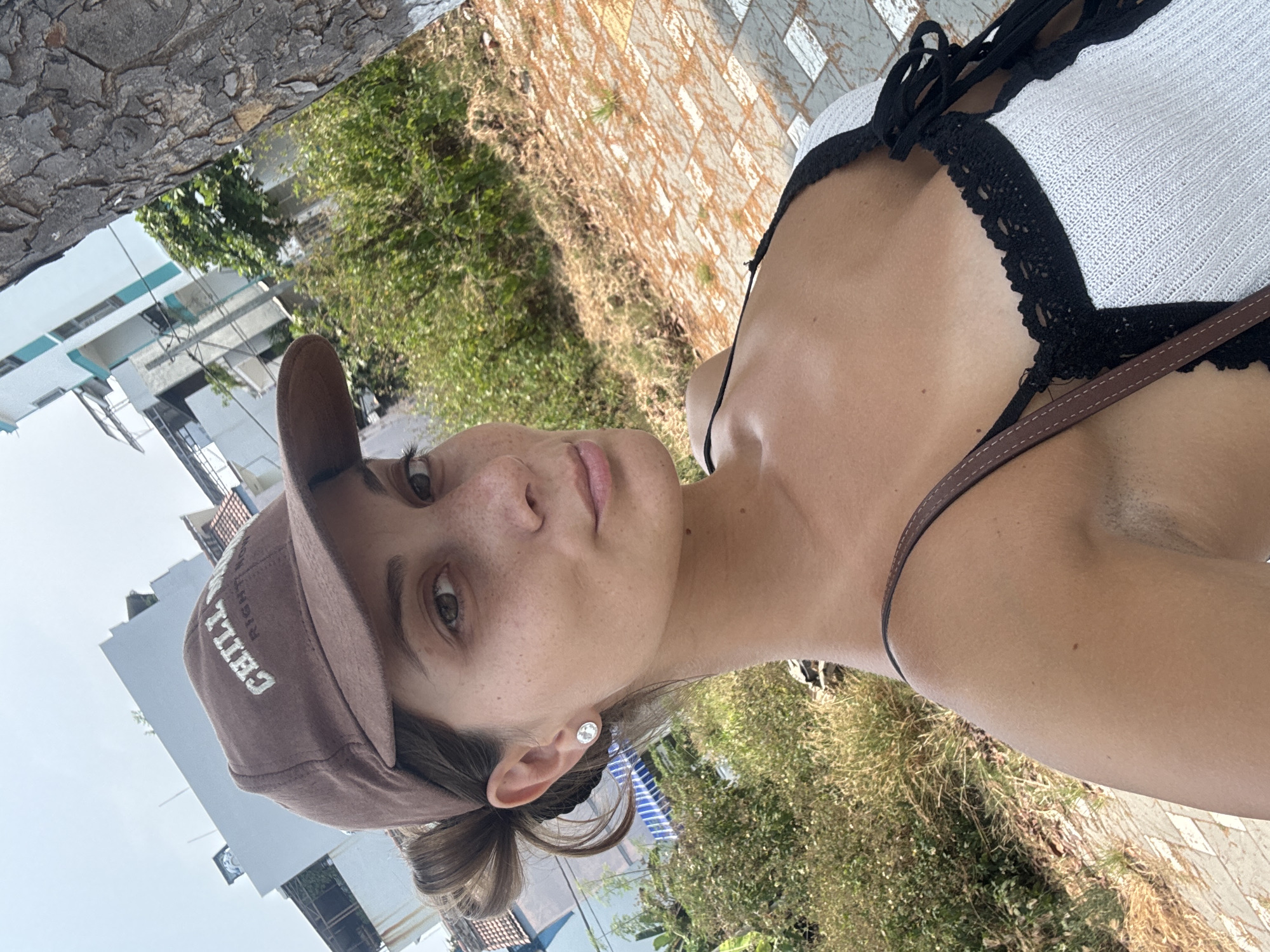 A woman is taking a selfie outdoors during daylight. She is wearing a brown baseball cap with white lettering, a white top with black lace trim, and a brown strap across her chest. She has light skin, freckles, and brown eyes. Her hair is pulled back in a low bun. She is wearing a small, round earring.
The background includes buildings, greenery, and a tiled walkway. Based on the architectural style, the photo could be taken in Đà Nẵng, Vietnam.