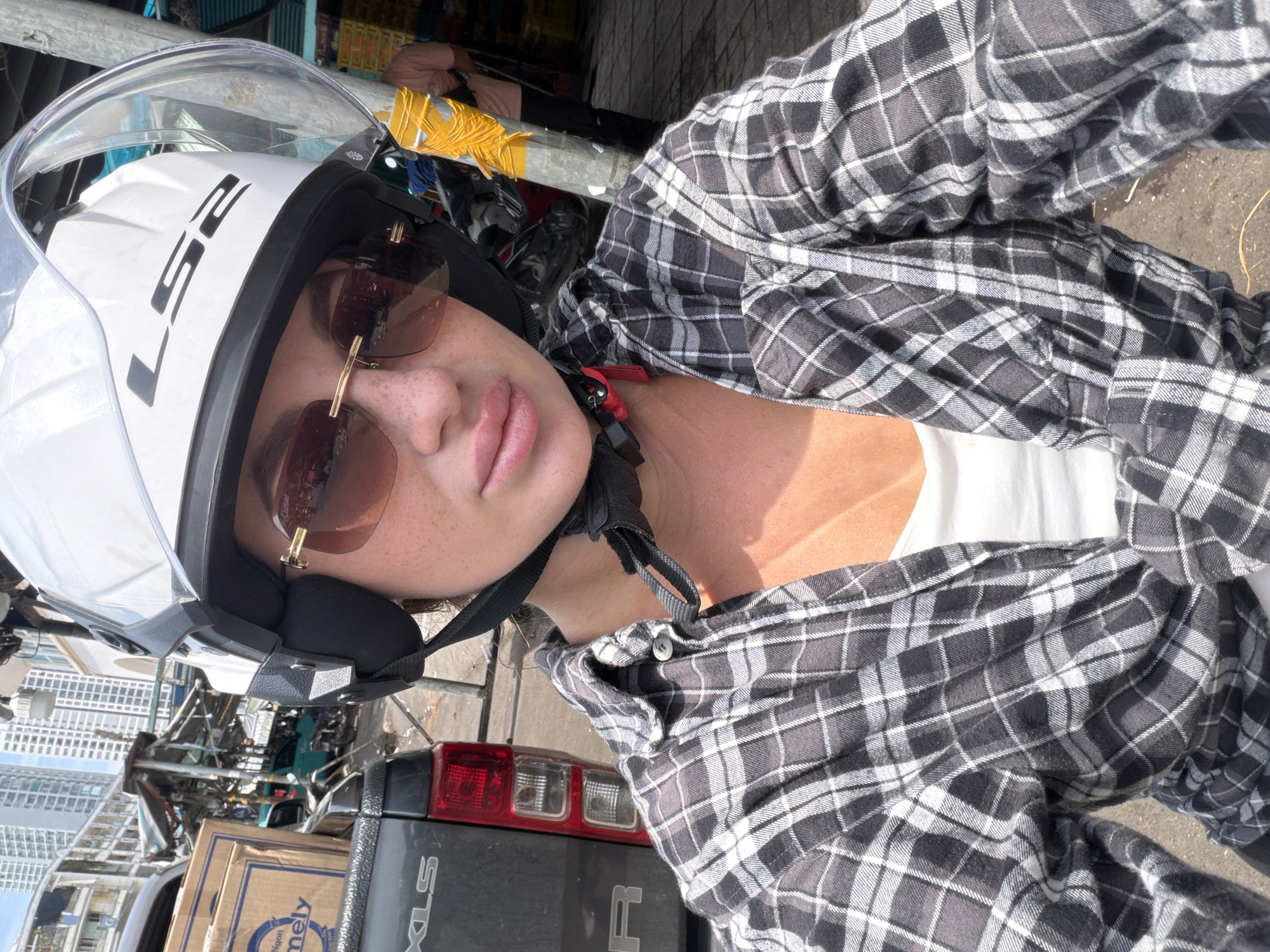 In Nha Trang Ward, Vietnam, a woman wearing a white LS2 helmet and sunglasses is taking a selfie on a motorcycle. She has a plaid shirt unbuttoned over a white tank top. In the background, a grey pickup truck is parked next to a market stall with various goods. Buildings and other vehicles are visible in the distance.