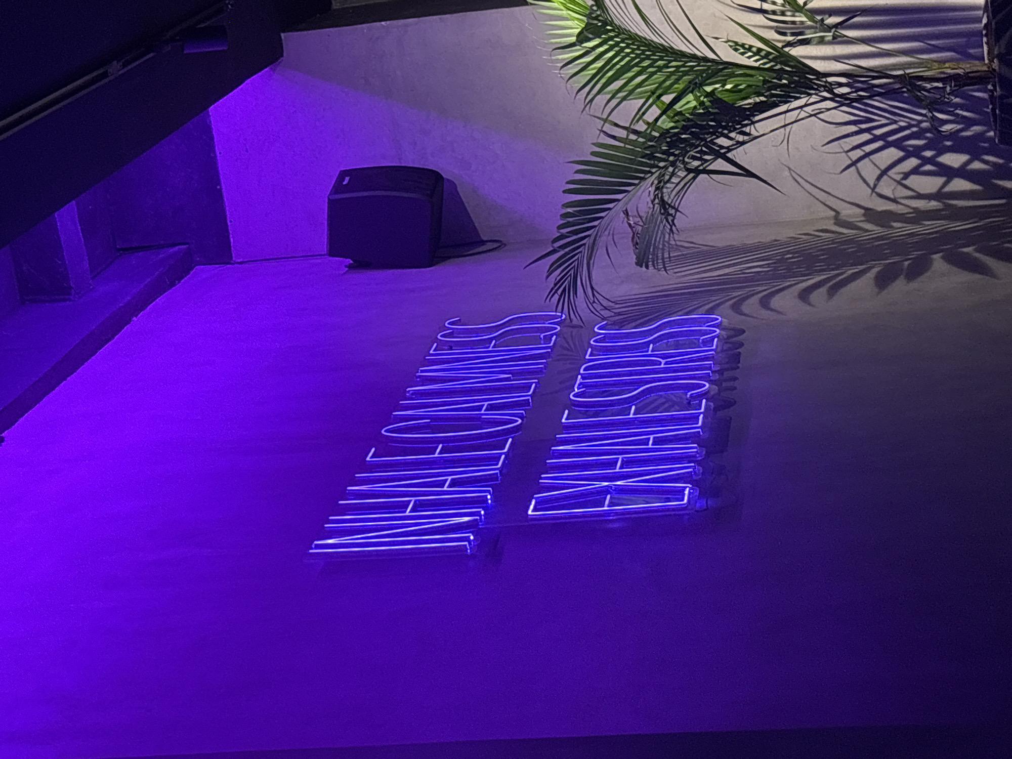 The image shows a partially obstructed neon sign in the Kuta Selatan area of Indonesia. The sign, emitting a blue-purple light, spells out "NINE GAMES WHITE SANDS". To the left, there is an indistinct dark structure that appears to be part of a building. Behind the sign and to the right, there is a potted palm casting shadows on the wall, suggesting a source of light other than the neon sign. A dark, rectangular object rests against the wall in the background. The floor is illuminated by the neon sign, contributing to an overall dark ambient light level. There are no people visible in the image.