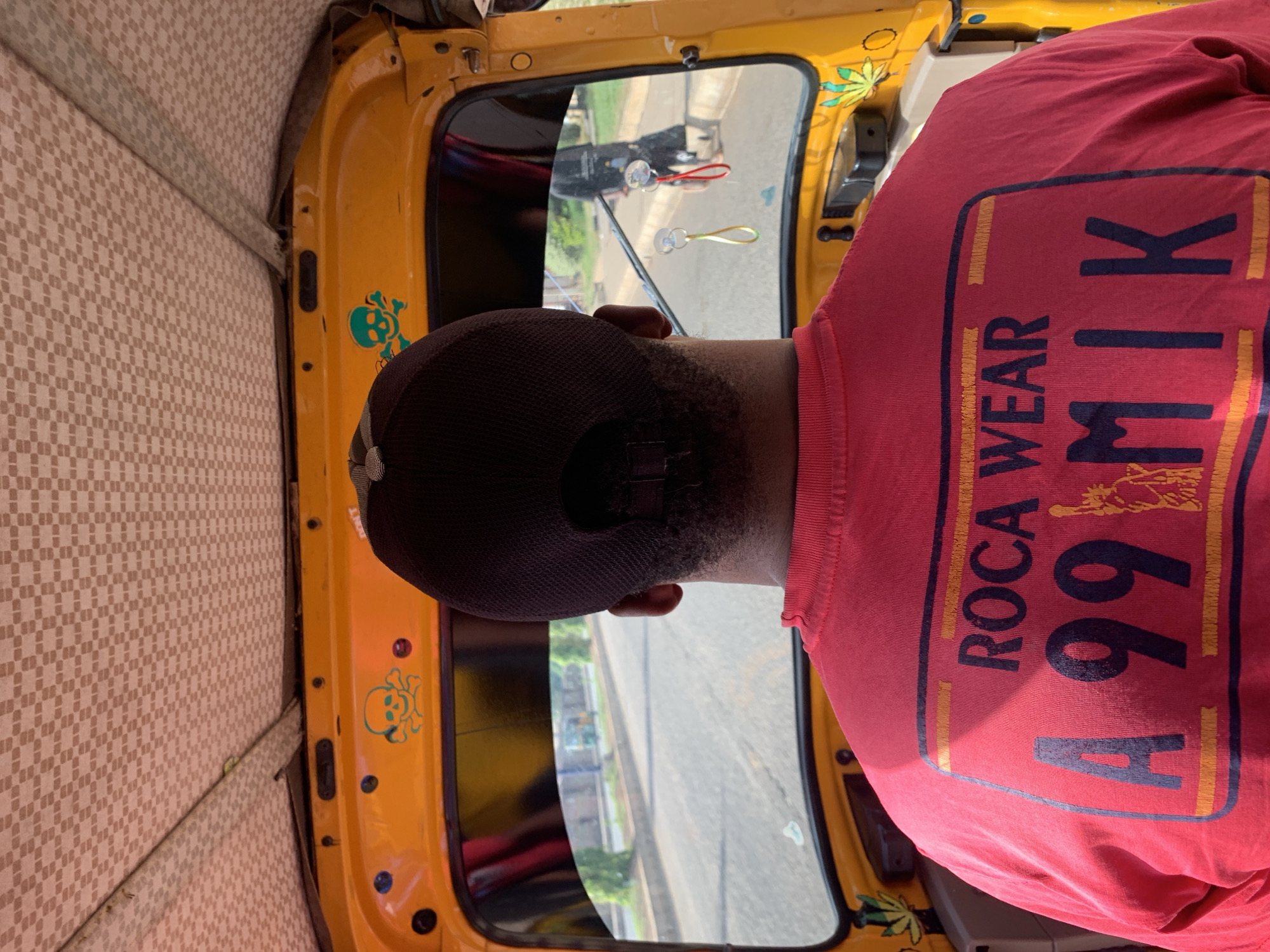 The image shows a person wearing a red t-shirt with "ROCA WEAR 499 MIK" printed on it, sitting inside a yellow vehicle, possibly a taxi or a tuk-tuk. The interior of the vehicle is visible, with a checkered pattern on the upholstery. Through the window, there's a view of a street and some buildings, suggesting an urban environment. Decorations, including a skull and a marijuana leaf design, can be seen on the yellow frame of the vehicle. The image appears to be taken from an elevated angle inside the vehicle. The street view implies that the location is Rafin Par, Nigeria.