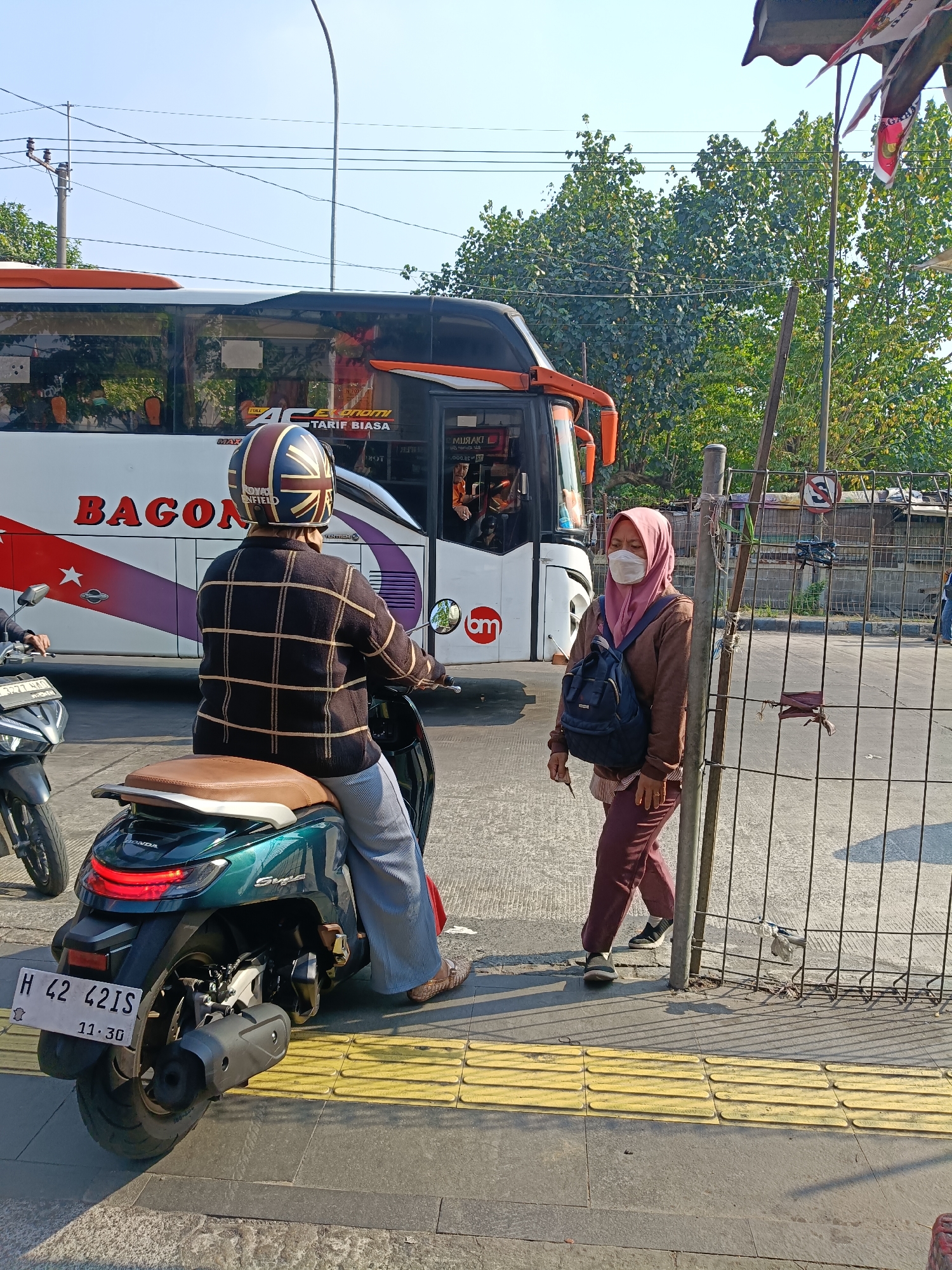 The image shows a street scene in Kedungrejo, Indonesia, on a sunny day. In the foreground, a person wearing a checkered sweater and a helmet with a Union Jack design is seated on a green scooter. They are wearing light blue pants and patterned shoes. To the right of the scooter, a woman wearing a pink headscarf, a brown jacket, and maroon pants is standing next to a metal fence. She is also wearing a white face mask and a dark blue backpack. Behind the scooter and the woman is a large white bus with orange trim, bearing the name "BAGON". The bus has tinted windows and appears to be in motion or stopped at a bus stop. The environment includes a sidewalk with yellow tactile paving, a street, and trees in the background. Overhead, power lines are visible against a clear blue sky. A second scooter is partially visible to the left of the main scooter. The scene depicts everyday urban life in Indonesia, possibly at a bus stop or intersection.