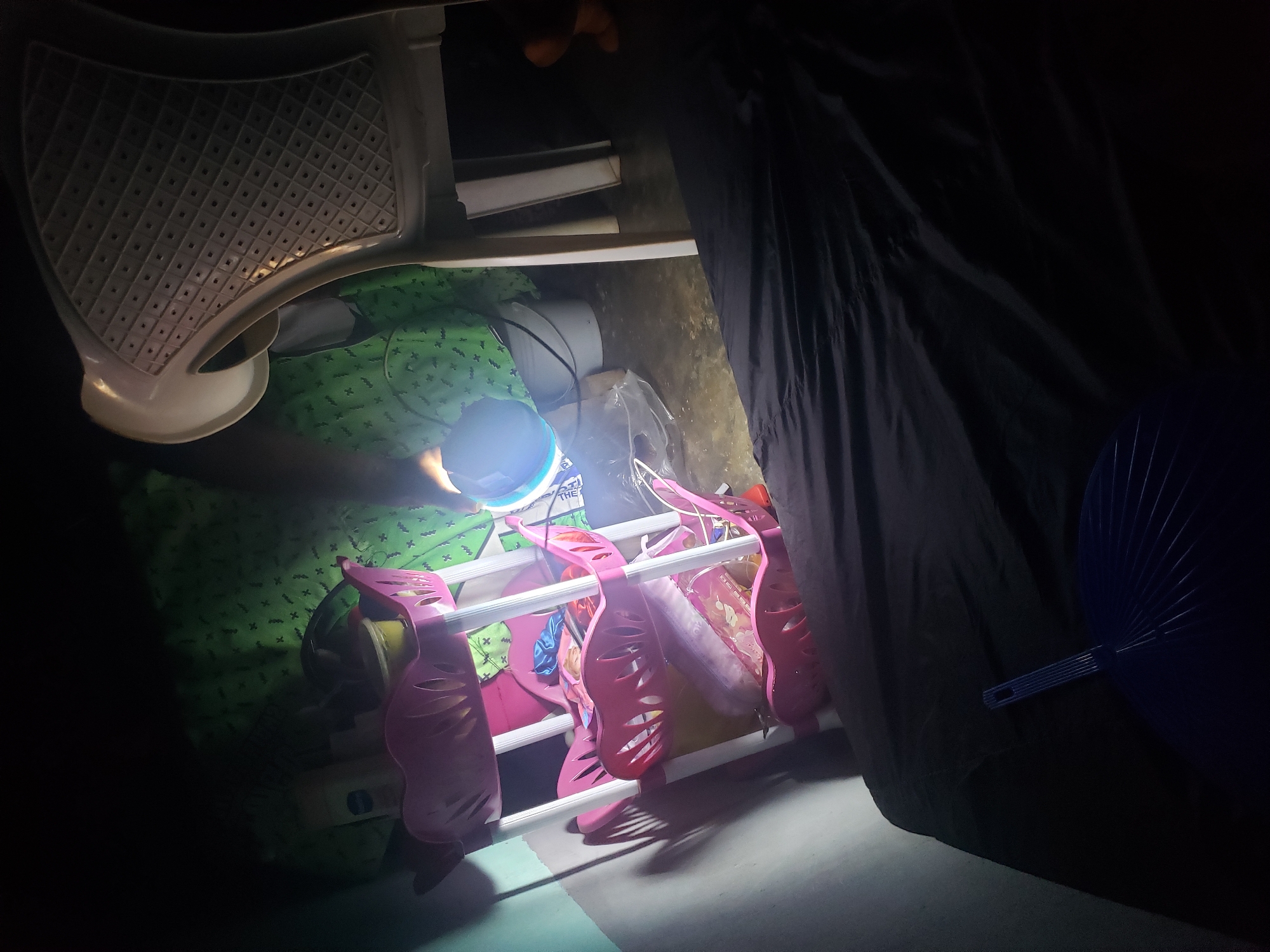 The image depicts a dimly lit interior scene, possibly a room, where a person is partially visible seated in a white plastic chair. The person appears to be holding a light source, casting a blue glow on a green patterned fabric in the background. In the foreground, a black fabric, possibly a bed or blanket, dominates the lower portion of the frame, with a blue foldable fan resting on it. To the left, a pink three-tiered plastic shelf unit holds various items, including bottles and containers. The environment suggests a basic living space. The provided location is the city of Kachia, Nigeria.