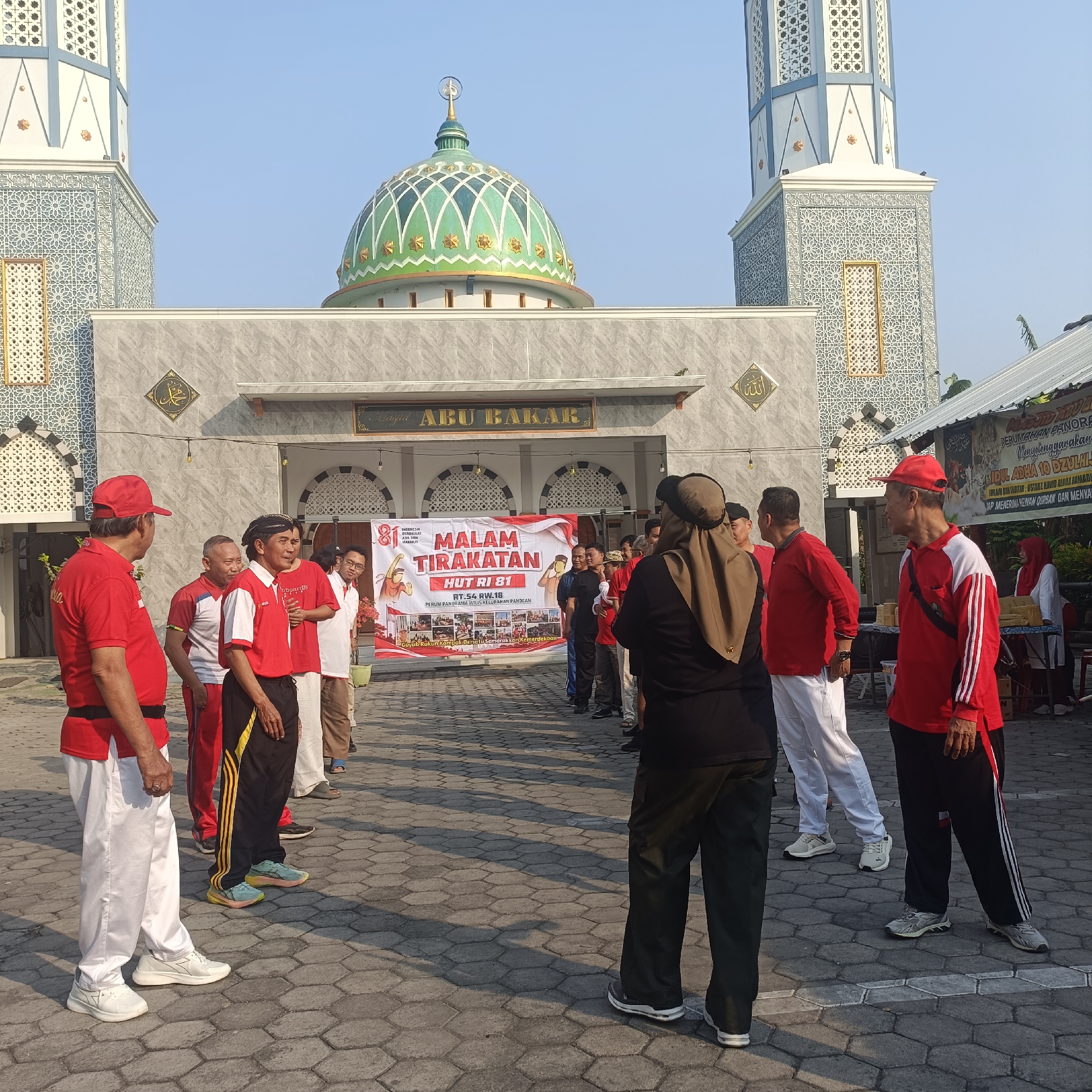 The image depicts an outdoor gathering in front of the Abu Bakar Mosque in Madiun, Indonesia. A group of people, predominantly men, are standing in what appears to be a courtyard paved with hexagonal tiles. Many of the men are wearing red shirts or tracksuits with white or black accents, and some are wearing red caps. In the foreground, two men in red shirts and white pants are facing each other, engaged in conversation. Behind them, a banner reads "MALAM TIRAKATAN HUT RI 81" indicating an event related to the 81st anniversary of Indonesian Independence. The mosque itself is visible in the background, featuring a green dome with white and gold ornamentation, and two tall minarets with intricate patterns. The sky is clear and blue, suggesting it is daytime.