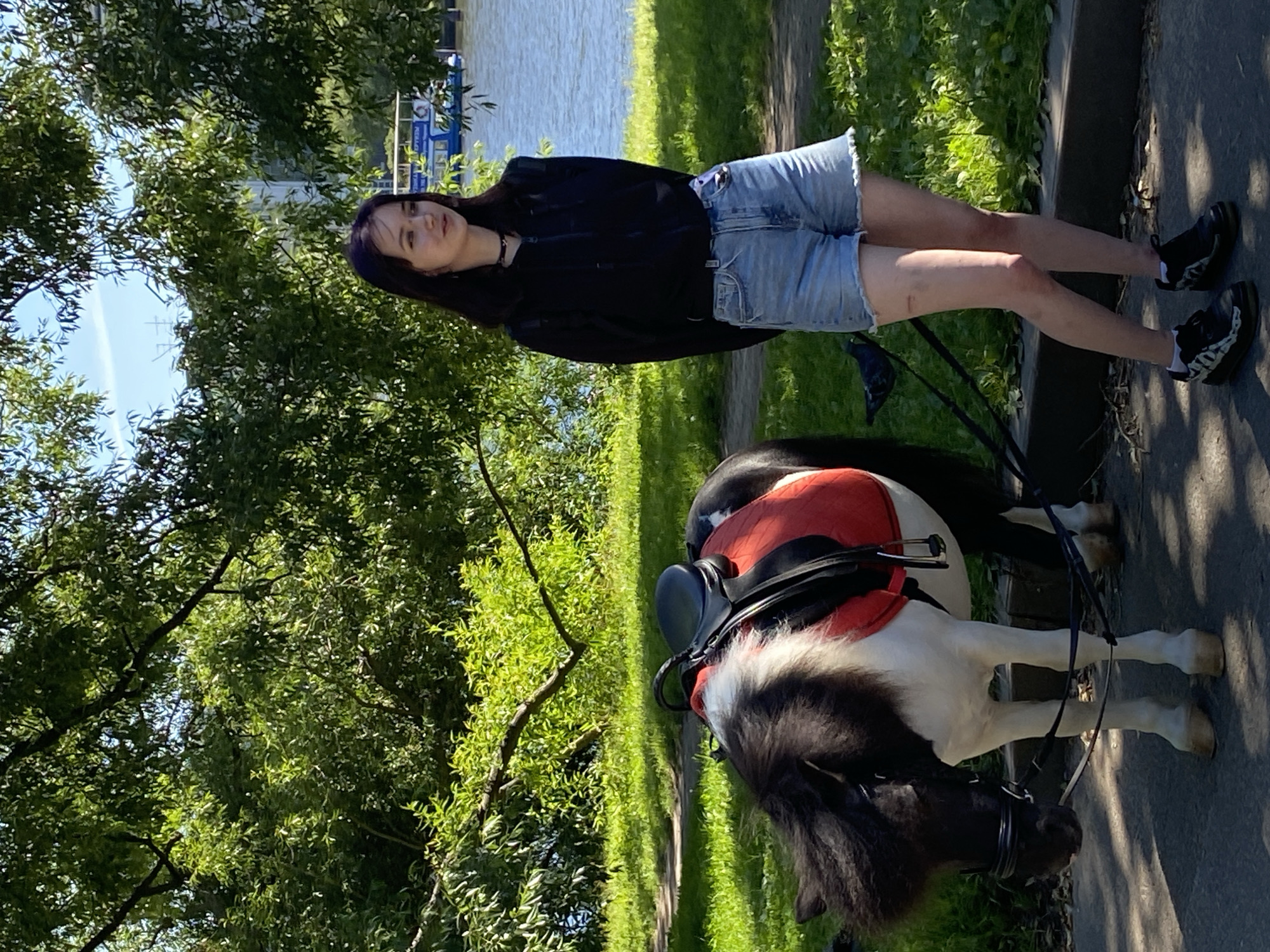 A young woman with dark hair stands next to a black and white pony in a park on a sunny day. The pony is equipped with a saddle and bridle, and appears to be tethered. The woman is wearing a black hoodie and denim shorts. Behind them, a body of water is visible, with a boat on the water. Lush green trees and grass surround the area, and sunlight filters through the leaves. The scene suggests a recreational setting, possibly in Saint Petersburg, Russia, given the presence of a boat on the water which could be a canal or river common in the city, and the overall park-like environment often found within urban areas of the city.
