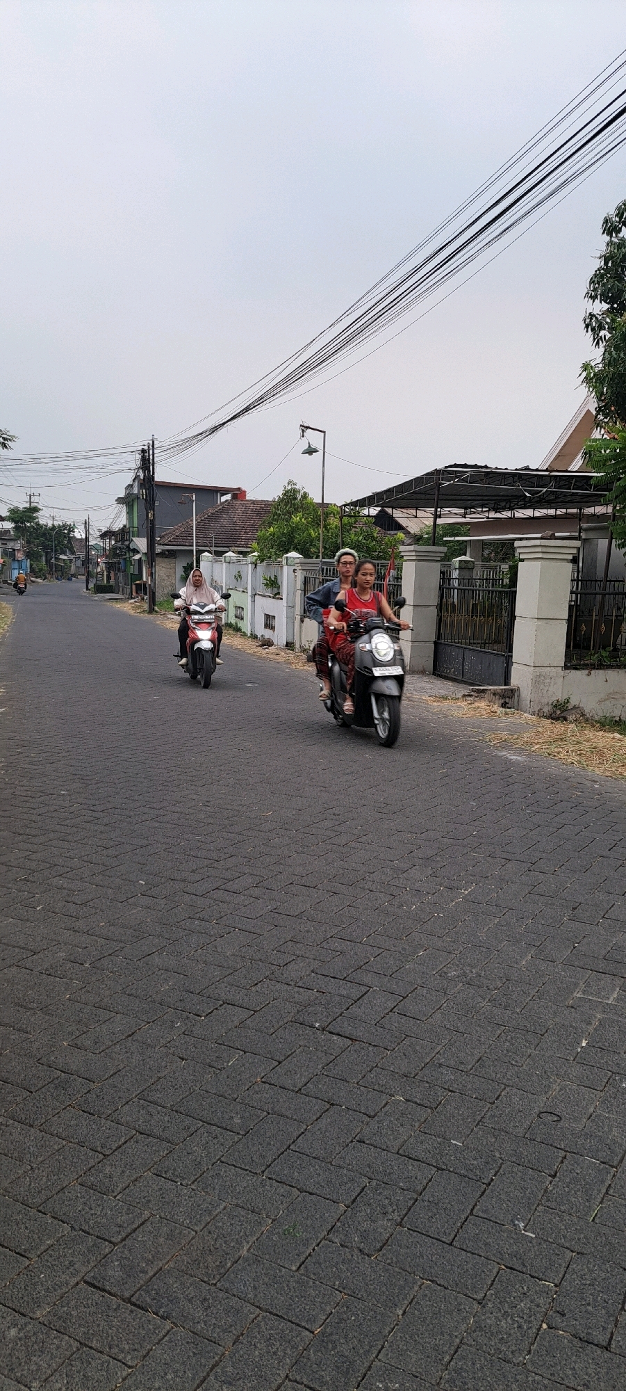 A low-angle, full shot captures a street scene in Ketanireng, Indonesia, under an overcast sky. The asphalt road is paved with dark grey interlocking bricks laid in a herringbone pattern. On the right side of the road, a white wall with a black gate marks the boundary of a property, beyond which is a covered patio with a tiled roof. Green trees are visible behind the wall and patio. Overhead, a dense network of black electrical wires stretches across the sky, supported by utility poles.
Two motorbikes are the central subjects. In the foreground, a woman in a red tank top and patterned pants rides a dark grey scooter. She is looking ahead and appears to be smiling. Seated behind her is a person with light-colored hair, wearing a denim jacket and dark pants, also looking forward.
Further down the street on the left, another motorbike, red and white, is ridden by a person wearing a light pink hijab and dark clothing. This rider is also looking forward. In the distance, more buildings and trees line the street, with a single distant motorbike visible. The overall atmosphere suggests a typical day in a residential area of a town.