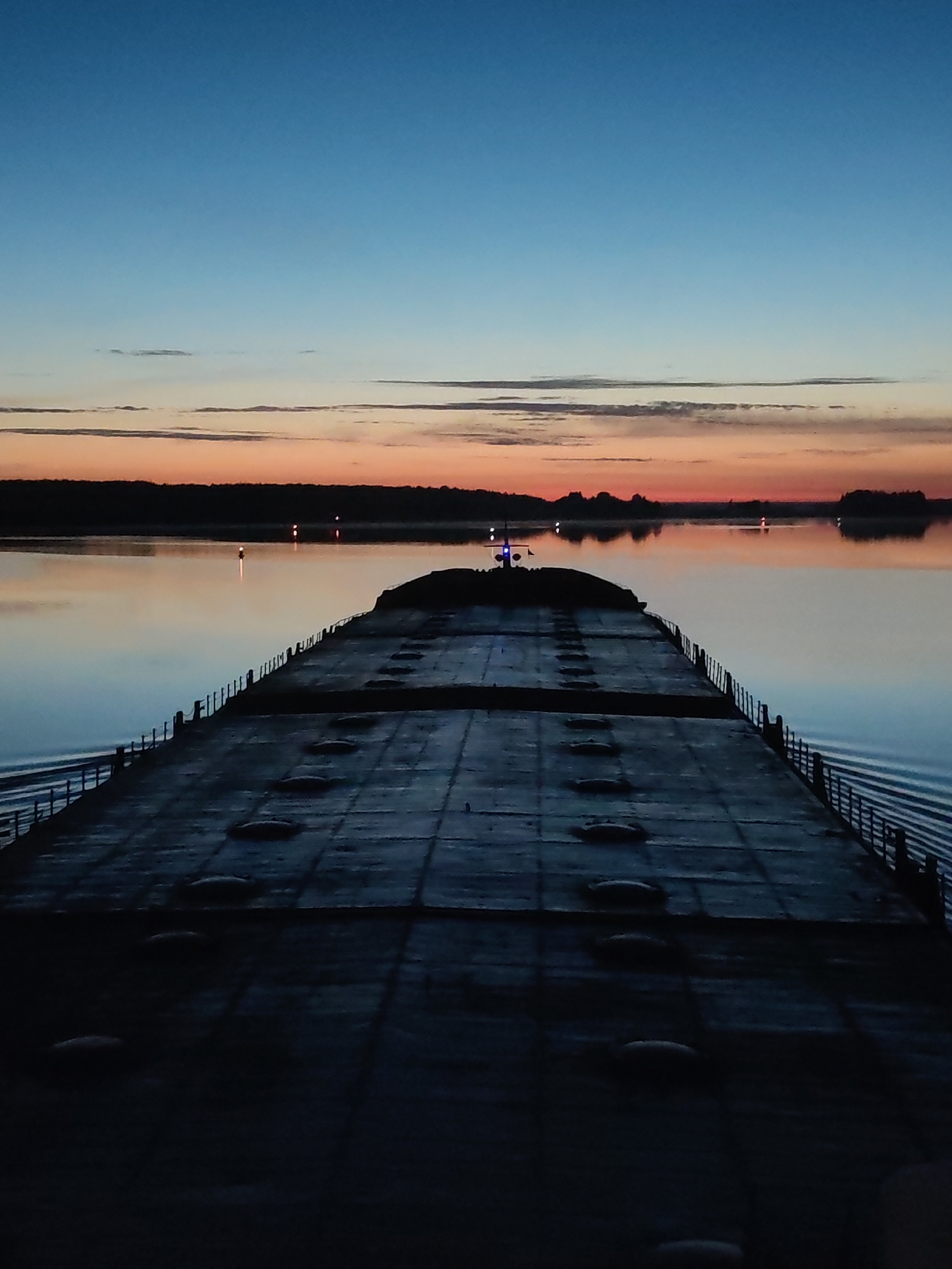 A long, dark barge dominates the foreground, its surface marked with hatches and framed by railings. The barge floats on calm water. In the distance, a dark treeline is silhouetted against a vibrant sunset with bands of orange and blue. Navigation lights punctuate the water's edge and center. The sky transitions from deep blue above to lighter hues near the horizon. The location is unknown.