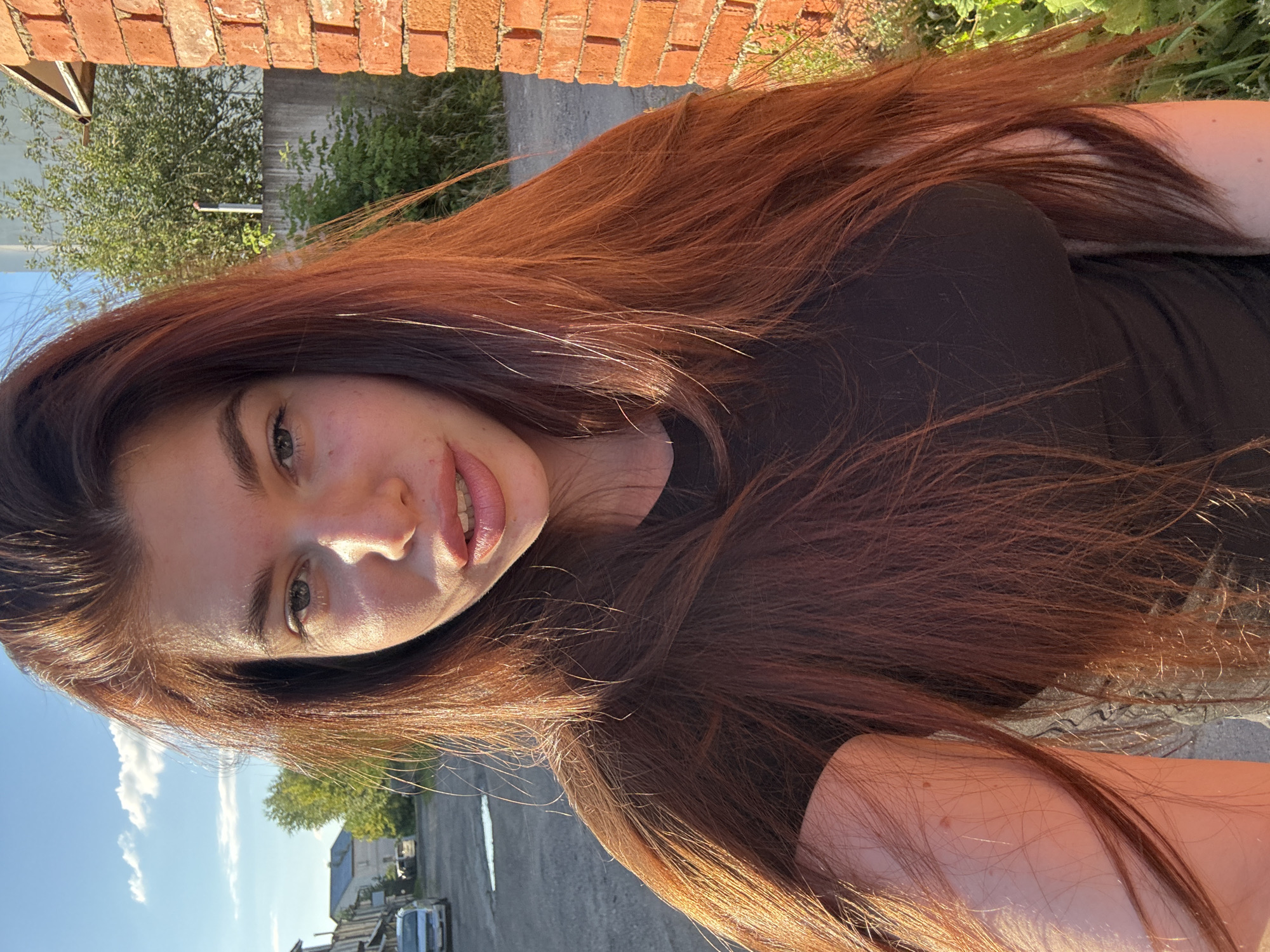 A medium shot portrait of a young woman with long, auburn hair and fair skin. She is outdoors, with a brick wall on the right side of the frame and a street with parked cars and buildings in the background. The sky is blue with scattered clouds. The woman is wearing a black t-shirt and is looking towards the camera with her mouth slightly open. Her skin is illuminated by golden hour lighting. The location is identified as Klin, Russia.