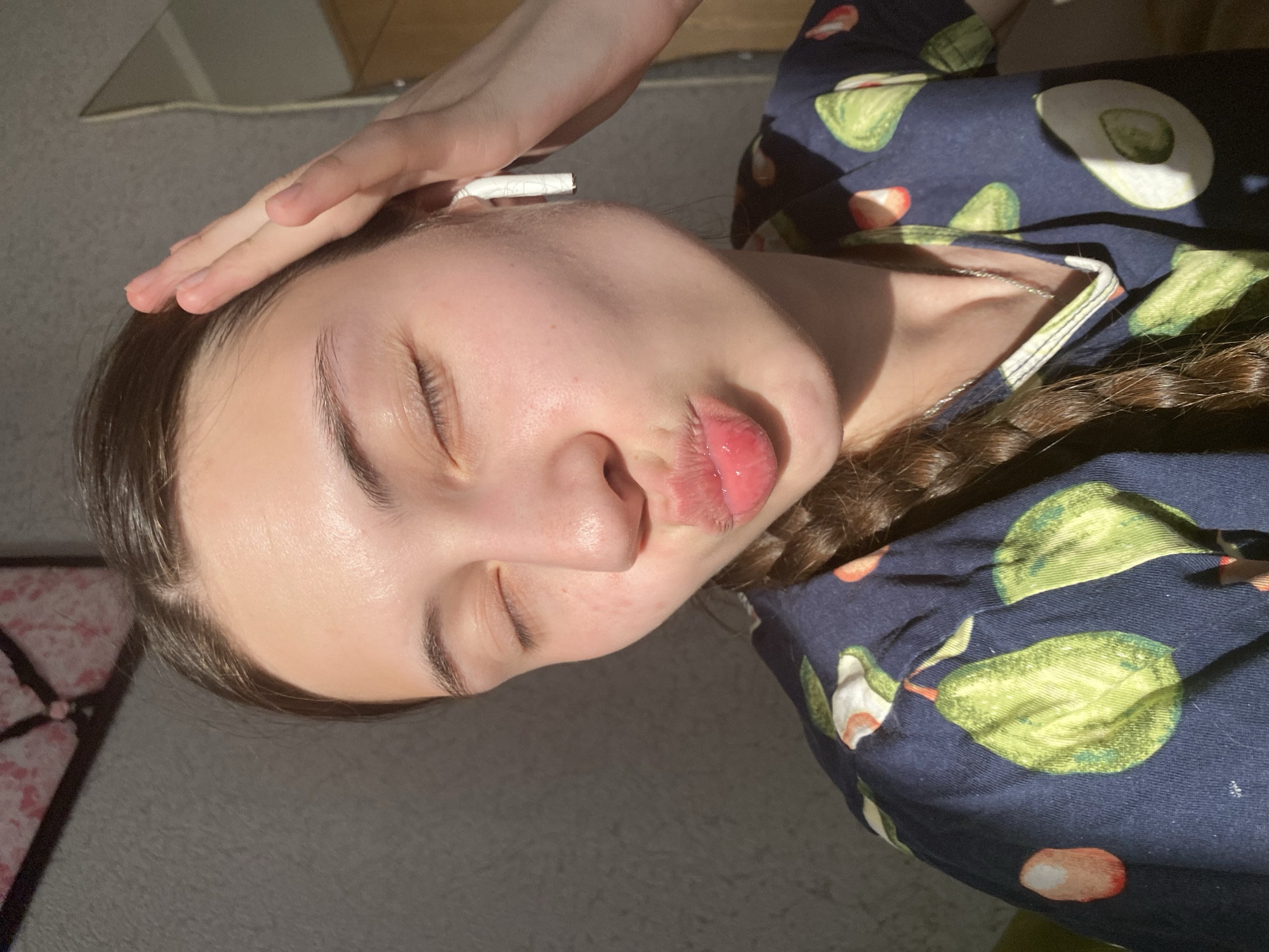 A young woman is positioned at an angle, lying down with her eyes closed. Her left hand rests on her forehead, and what appears to be a white cigarette is placed near her ear. She has a single braid that is visible on her right side. She is wearing a blue garment printed with avocados and strawberries. The background is a carpeted floor and a wardrobe or cabinet in a blurred background. This scene takes place indoors, but no information indicates this is in the city of Votkinsk, Russia.