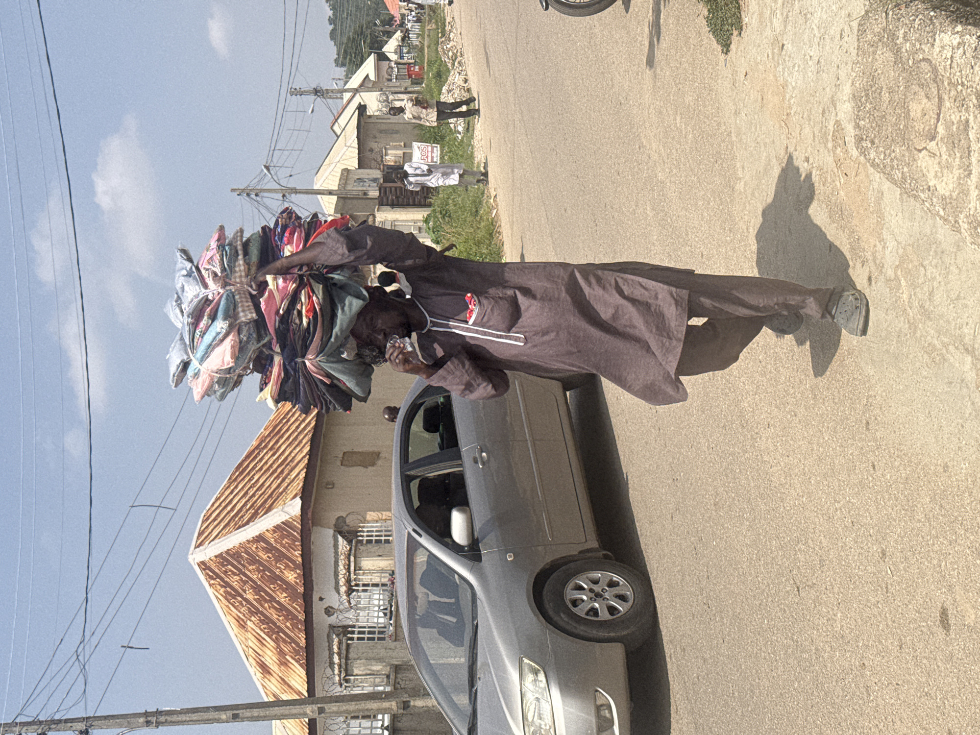 In Abuja, Nigeria, a man walks down a paved road, carrying a large, colorful stack of fabrics balanced on his head. He is wearing a long, dark brown tunic and pants, with dark footwear. He holds a clear plastic cup in his right hand, appearing to drink from it. A silver sedan is parked on the left side of the road, and in the background, buildings with corrugated metal roofs are visible. The sky is a light blue with scattered clouds. Several other people are seen walking in the distance. The environment appears to be an urban street with power lines overhead.
