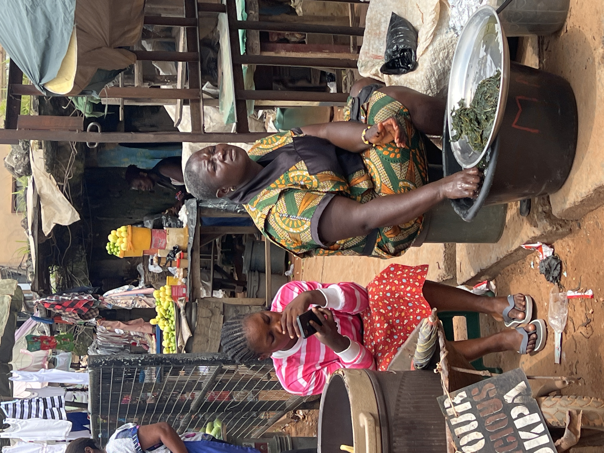 This media file captures a scene in Nkpor, Nigeria, likely during the daytime. The primary subjects are two individuals and a bustling market environment.
In the foreground, a young girl with braided hair is seated and engrossed in her smartphone. She is wearing a pink and white striped long-sleeved shirt and a red dress with white polka dots. To her right, resting her feet on a small stool, she wears grey flip-flops.
Behind and slightly to the left of the girl is an adult woman. She appears to be a vendor, seated on a low stool and tending to a large basin filled with green leafy vegetables. She is wearing a vibrant, patterned dashiki-style dress in green, yellow, and orange, and a dark headscarf. Her expression suggests she might be tired or in discomfort. She is using a metal bowl to scoop the greens from a larger bucket.
The setting is an outdoor market stall, characterized by makeshift structures made of wood and metal. Shelving units are laden with goods, including stacks of clothing and yellow bunches of what appear to be bananas or plantains. Laundry hangs on a clothesline in the background. The ground is dirt.
Notable details include the presence of plastic bags, one black bag near the woman and another lighter-colored bag further in the background, suggesting ongoing market activities. The lighting indicates it is daytime, and the overall atmosphere appears to be that of a busy, possibly impoverished, market. A sign with white lettering on a dark background is visible at the bottom, but the text is not fully legible.