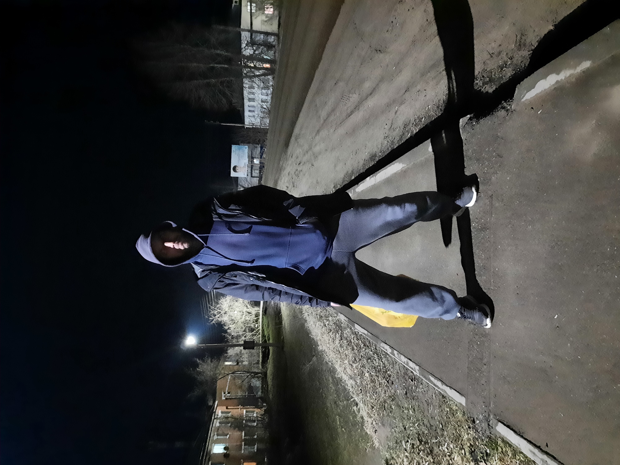 A person wearing a blue hoodie, dark pants, and black shoes is standing on a paved path next to a curb and a strip of overgrown grass in what appears to be an urban area at night. The scene is illuminated by a street light in the background, casting a long shadow of the person onto the pavement. Behind the person, there are several buildings and bare trees. The image was taken in Krasnogorskoye, Russia.