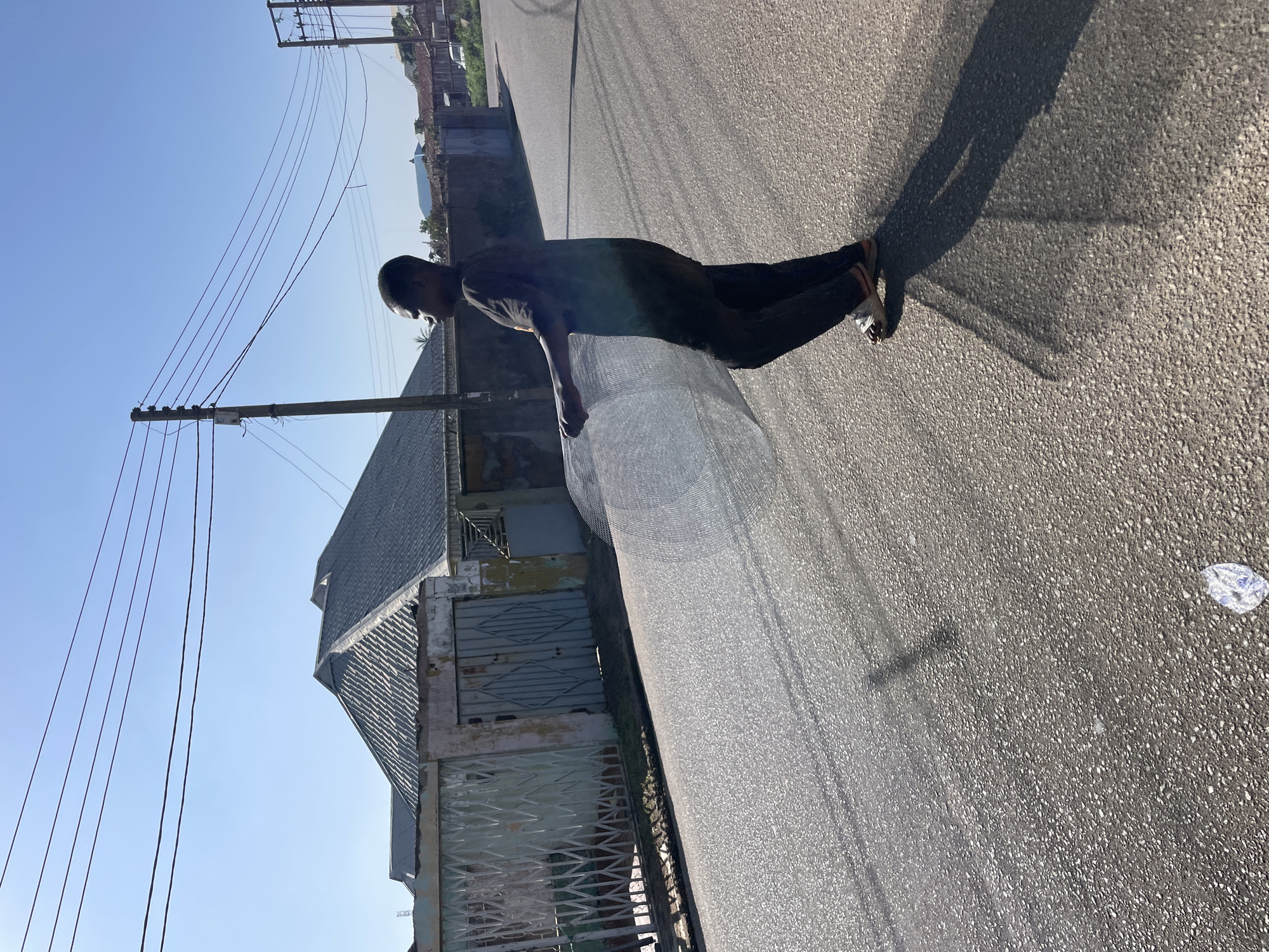 A young person is walking on a paved road in Jos, Nigeria, carrying a large, rolled-up piece of netting. The person is wearing a dark t-shirt and dark pants with sandals. They are walking from left to right in the frame, with their body angled away from the camera. The netting is held in front of them, obscuring part of their torso. The road is asphalt and appears to be in good condition, with visible tire marks and shadows cast by overhead power lines. The surroundings include a fence with a gate to the left of the road, and houses with corrugated metal roofs in the background. Overhead power lines stretch across the sky, supported by utility poles. The sky is clear and blue, indicating a sunny day. A discarded plastic wrapper is visible on the road in the lower left portion of the image.