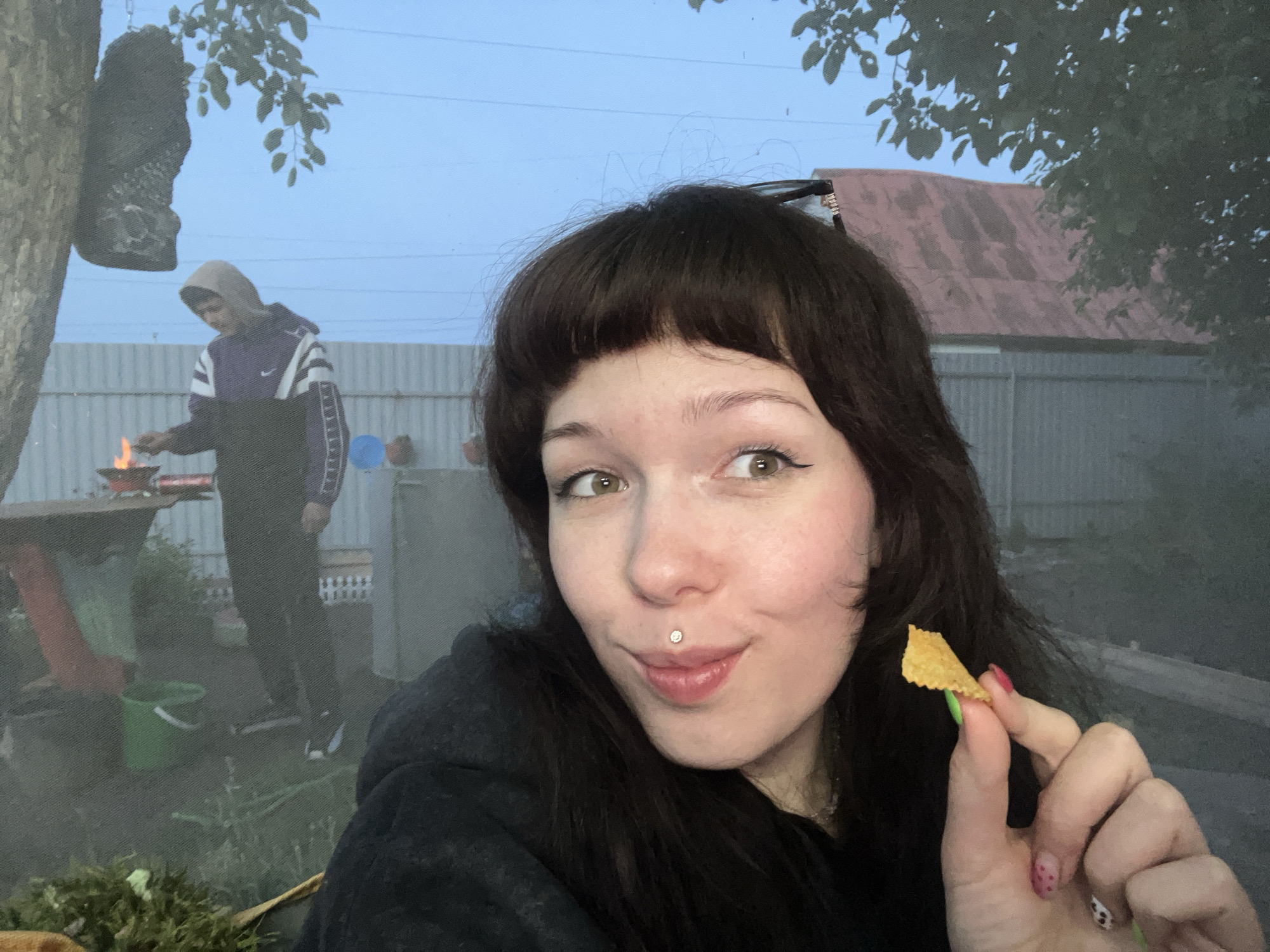 The scene is set outdoors, depicted as taking place in Chaikovsky, Russia, during what appears to be dusk or early evening under a light blue sky. In the foreground, a young adult female with dark brown hair and bangs smiles slightly towards the viewer. She has green/hazel eyes, prominent eyeliner, and a silver stud labret piercing above her upper lip. She is wearing a dark-colored top, possibly a hoodie, and holds a single, golden-brown, triangular chip between the thumb and index finger of her right hand. Her fingernails are visibly painted with multi-colored designs, including bright green, red with white dots, and white with black cow print.
In the mid-ground, to the left, a male individual is visible. He wears a light-colored hoodie pulled over his head, with a purple and white striped long-sleeved top featuring a white Nike swoosh logo on the chest. He is standing near an elevated metal fire pit or brazier, actively tending to a visible fire within it. A bright green bucket with a white handle is positioned on the ground near his feet, to the left of the fire pit.
The background features a corrugated metal fence stretching horizontally, behind which a building with a weathered, reddish-brown corrugated metal roof is visible on the right. A thick tree trunk stands on the far left, from which a dark, irregular object is suspended by a chain. Green foliage from tree branches extends into the upper left and right portions of the frame. The ground appears to be a mix of dirt and sparse vegetation, with a generally hazy atmosphere, likely due to smoke from the fire.
