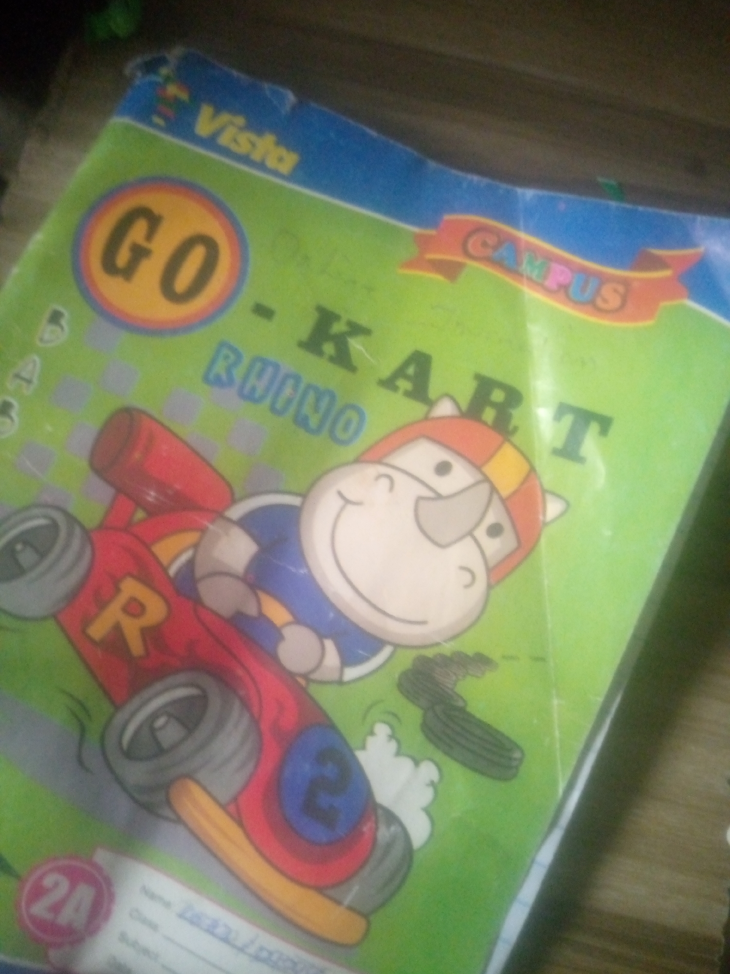 A close-up, slightly angled, blurry shot shows the cover of a notebook. The notebook is green with a blue border. The title of the notebook is "GO - KART RHINO" in large black letters. There is a cartoon of a rhinoceros wearing a racing helmet driving a red race car. The rhinoceros has a cheerful expression. The notebook also features the words "Vista" and "CAMPUS" on the blue border. In the bottom left corner of the notebook, there is a circular label with "2A" inside. Below the illustration, there is a section with lines for "Name," "Class," and "Subject," with some illegible writing. The notebook is placed on a wooden surface, possibly a table or desk. The lighting is dim, and the image quality is poor, with a lot of grain. There are no people visible in the image.