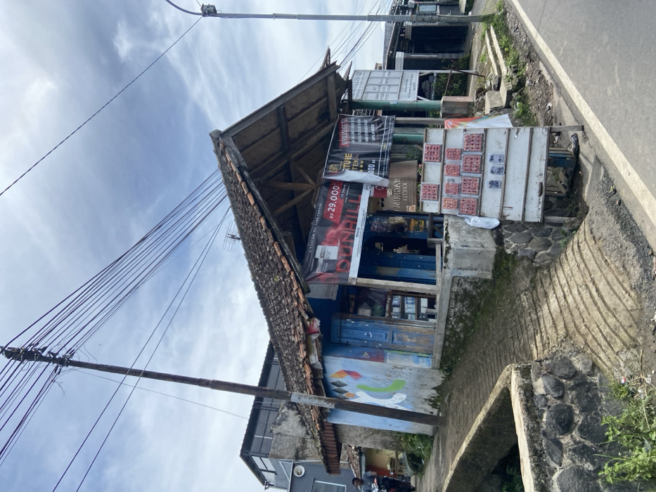 The image depicts a small, weathered wooden structure with a corrugated metal roof, located in the village of Rancabali, Indonesia, at the coordinates -7.118 latitude, 107.435 longitude. The building has a blue and white painted exterior, and is adorned with various signage and advertisements. A person is standing outside the building, appearing to be interacting with a sign displaying red goods. The structure sits on a slight incline, with a stone ramp leading up to it, and a stone wall bordering a small ditch in the foreground. The scene is set against a backdrop of a paved road and a cloudy sky, with numerous power lines overhead. The building is surrounded by vegetation, including a few trees.
