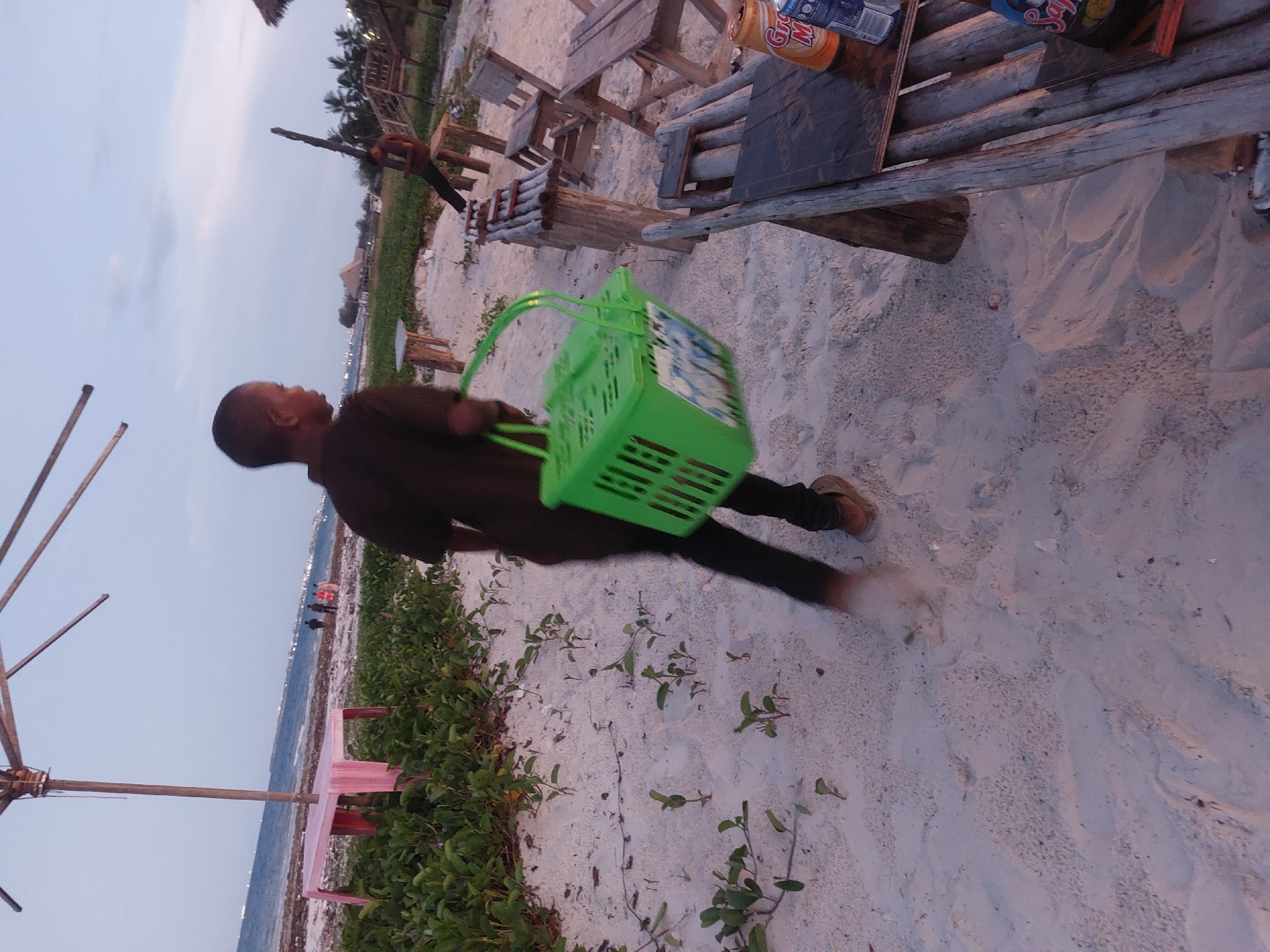 A young boy is running on a sandy beach. He is holding a green plastic basket. The beach is littered with vegetation and wooden structures that appear to be tables and chairs. In the background, the ocean and a shoreline dotted with buildings are visible. The photo was taken in Dar es-Salaam, Tanzania.