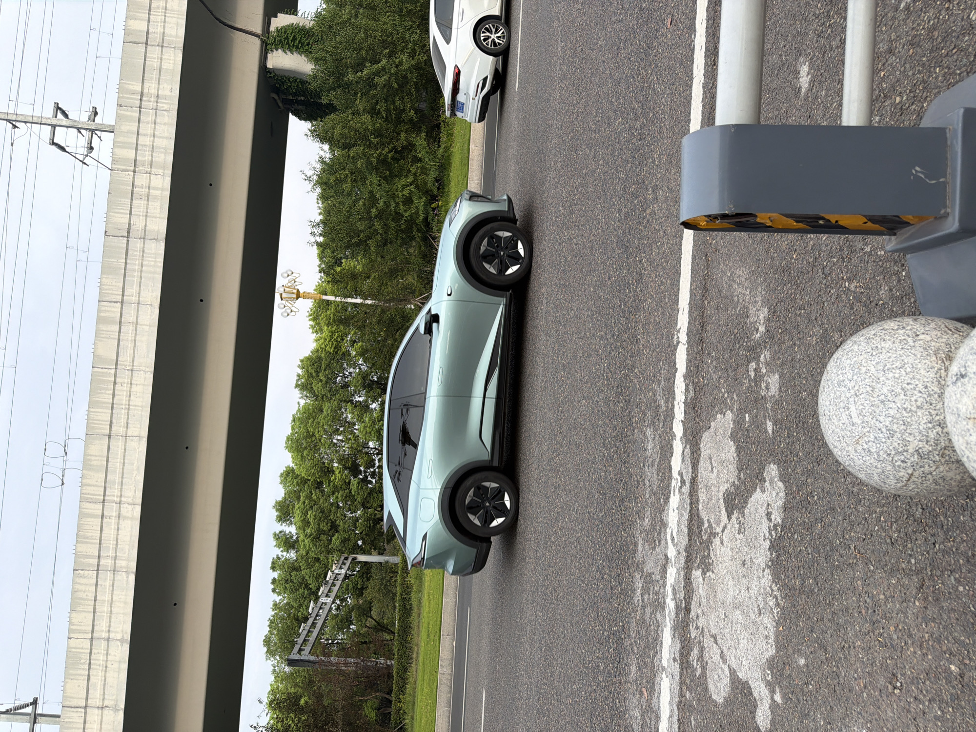 In Jiangjin, China, a pale teal electric vehicle is driving on a multi-lane road under a concrete overpass. The overpass has overhead power lines and a street light with multiple lamps. The electric car is a coupe-style SUV with black trim and distinctive black and silver wheels. To the right of the electric car, a white sedan is also traveling on the road. The road surface is asphalt with white lane markings. In the foreground, a gray guardrail is visible, along with two round, granite-like bollards. Lush green trees and vegetation line the side of the road. The sky is overcast.