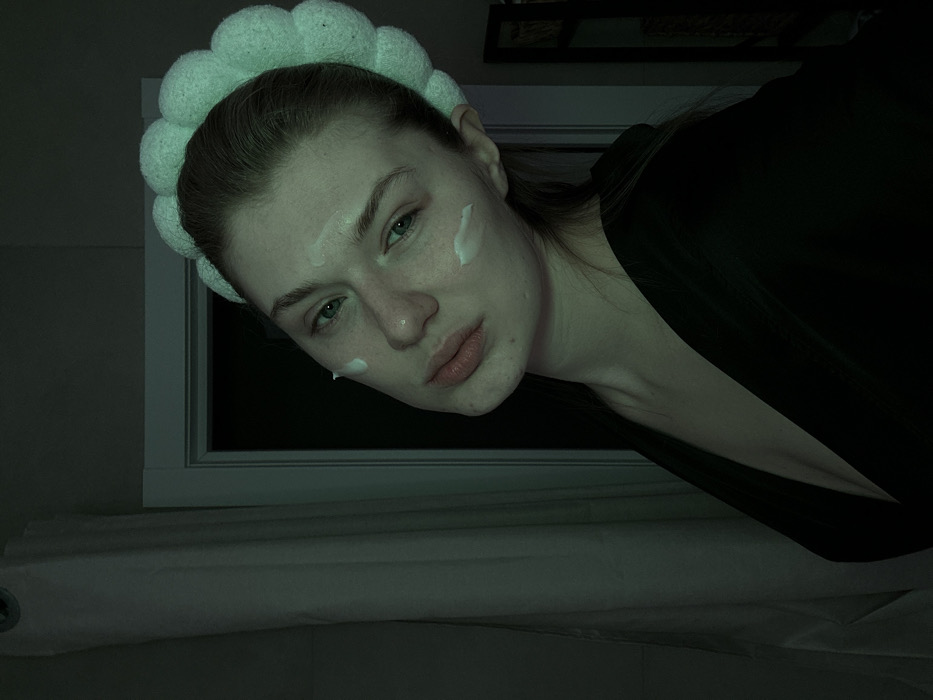 A woman with fair skin is angled to the viewer, donning a puffy, light-green headband. Dots of white cream are visible on her face. She has light-green eyes and pursed lips. She wears a dark, low-cut garment. Behind her is a white-framed window with a dark backdrop and a white, rolled-up object on what seems like a bed. The lighting is low, giving the image a muted tone.