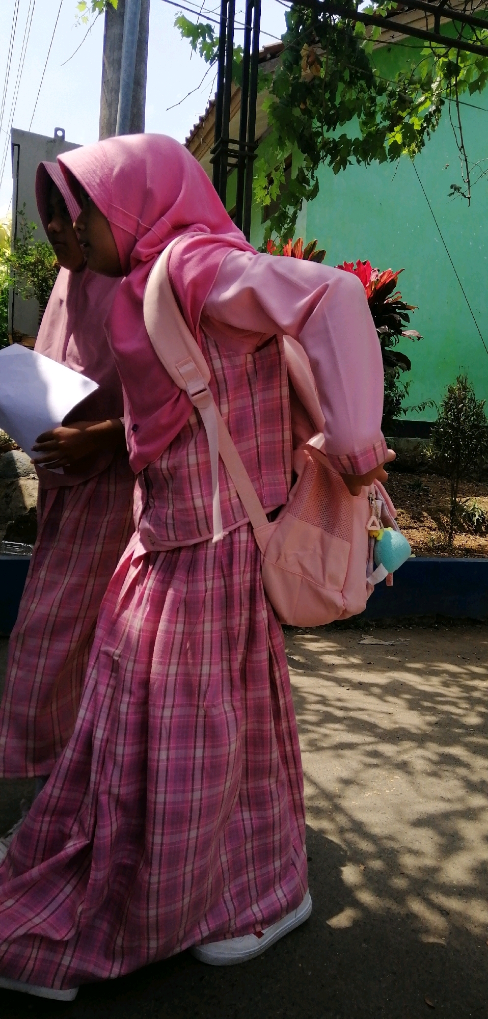 Two young females wearing matching pink hijabs and plaid dresses, likely students, are walking on a paved area. One is holding a white document. The individual in the foreground has a pink backpack slung over her shoulder. The background features a green wall, some foliage, and a utility pole. The scene appears to be outdoors, under sunlight, with shadows cast on the ground. The location is identified as Ajibarang, Indonesia.