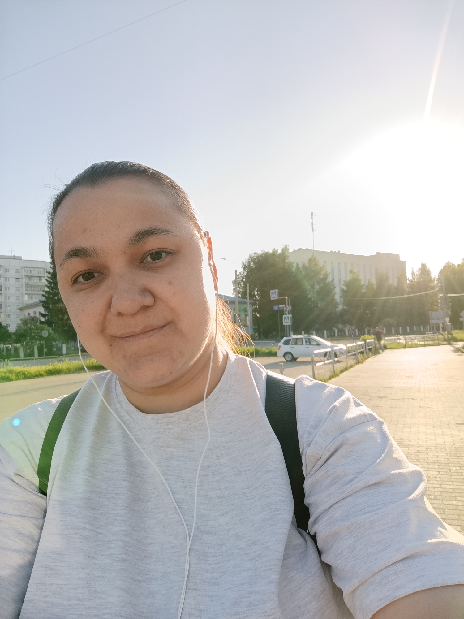 A woman with dark hair pulled back and wearing a gray long-sleeved shirt with a green backpack strap is taking a selfie outdoors in what appears to be the city of Chaikovsky, Russia. She has earphones in her ears. Behind her is a sidewalk, a street with a white car, buildings, trees, and traffic signs. The bright sunlight suggests it is daytime.