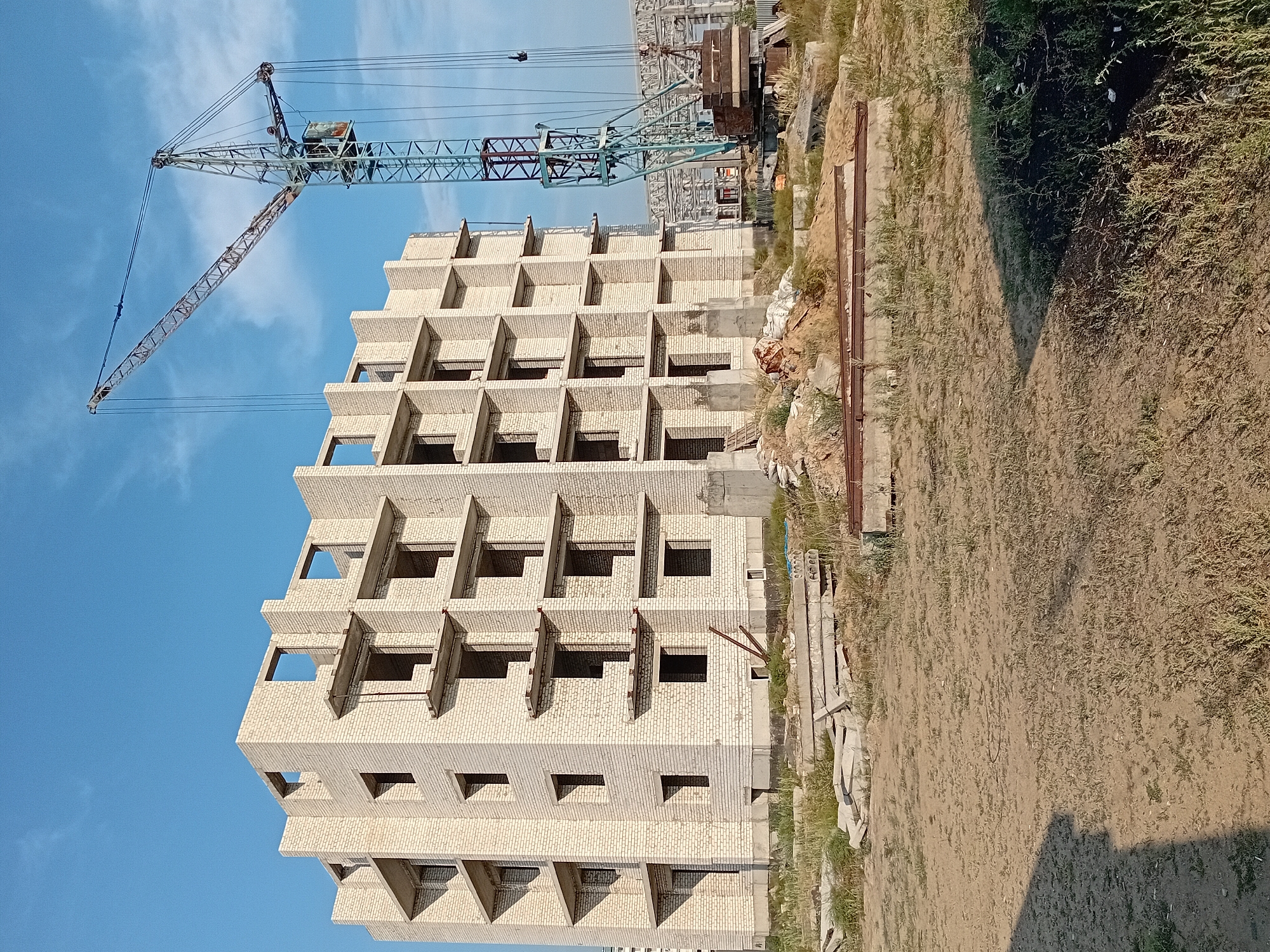 The image depicts an outdoor construction site in the city of Semey, Kazakhstan, under a clear blue sky with some wispy clouds. A multi-story building made of white bricks, currently in its skeletal phase with open window frames and unfinished concrete balconies, dominates the left side of the frame. The building appears to be a residential or commercial structure under construction.
To the right of the building stands a tall construction crane, its boom extended over the site, indicating ongoing work. In the foreground and around the base of the building, there are piles of construction debris, sand, concrete slabs, and other building materials, suggesting an active but not necessarily organized, work environment. The ground is a mixture of dirt and sparse, dry grass. In the background, more structures, possibly also under construction or part of an industrial complex, are visible. The overall impression is one of urban development or renovation in progress.
