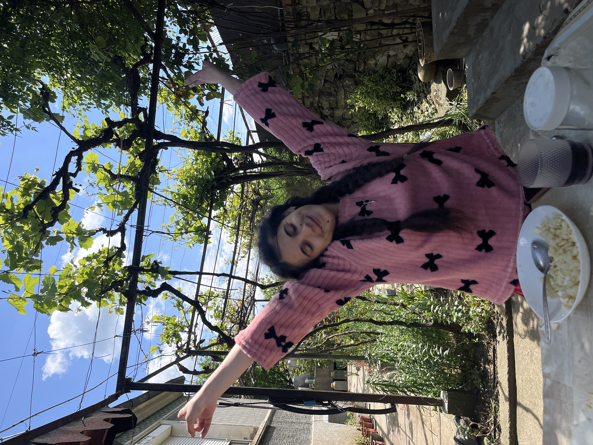 A young woman with long dark hair, possibly braided, is lying on a ledge in Lozarevo, Bulgaria. She is wearing a pink bathrobe with black bows. She has her arms outstretched. Above her is a metal framework covered in grape vines with green leaves. The sky is visible through the vines. To the right of the woman is a concrete ledge with a plate of food and some cups and bottles. Behind the ledge is a stone wall covered in vegetation. Below the woman and to the left is a building with a gray exterior.