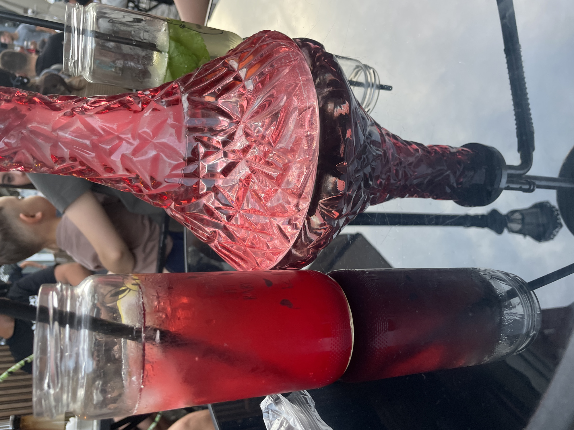 This is a medium close-up, eye-level, outdoor shot of a table with two drinks and a hookah on it, set against a blurred background of people. The drinks appear to be beverages in glass jars, with one on the left being a deep red color and the other on the right being a lighter, greenish hue with mint leaves. A red, patterned glass hookah base is positioned prominently in the foreground, between the two drinks. The table's surface is reflective, showing a clear reflection of the hookah base and the sky. The background is filled with the silhouettes of people, suggesting an outdoor social gathering or restaurant setting. The lighting is natural and appears to be daylight.