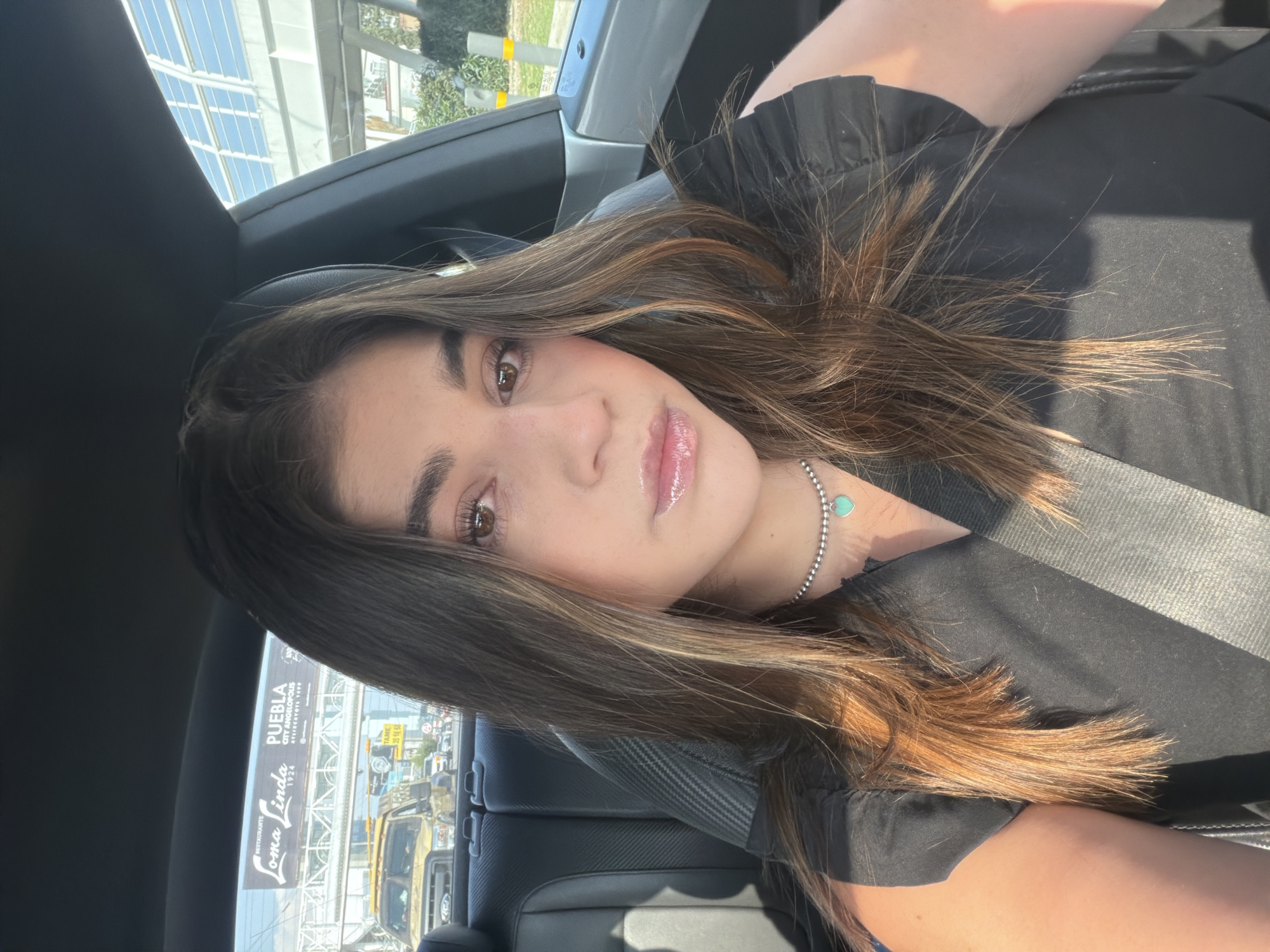 A woman is seated in the driver's seat of a car. She is wearing a black top, and a silver necklace with a turquoise heart-shaped pendant. A seatbelt is visible across her chest. The background shows a street scene with vehicles and a sign that reads "Loma Linda Restaurante, Puebla, City Angelopolis." This indicates that the location is likely San Bernardino Tlaxcalancingo, Mexico, which is near Puebla City.