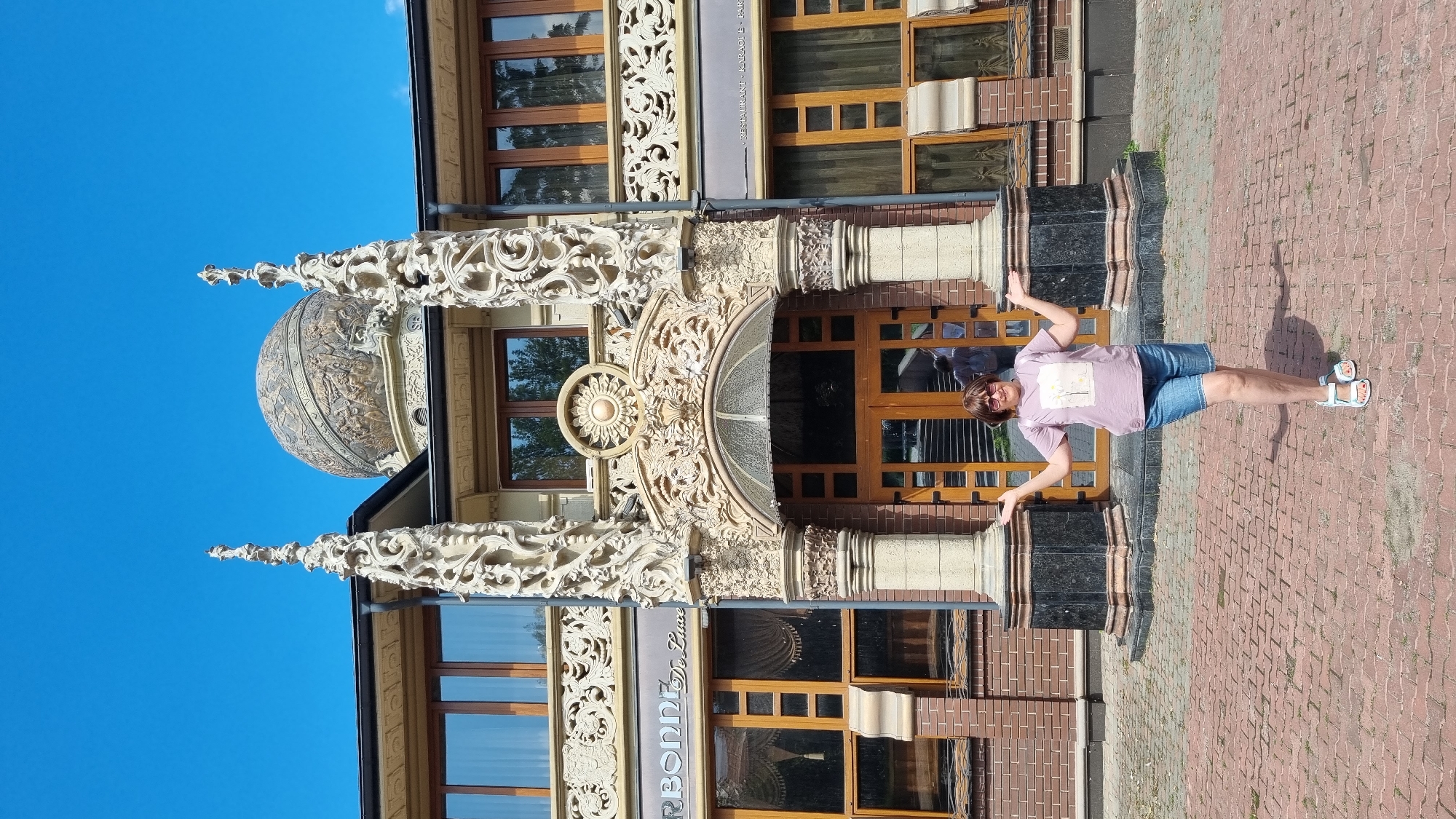 A woman in a pink t-shirt and denim shorts stands with her arms outstretched in front of a building with ornate architectural details. The building features intricate carvings, a large dome, and several windows. The woman is wearing white sandals and sunglasses. The ground in front of the building is paved with red bricks. The sky is clear and blue. The building is located in Chernivtsi, Ukraine, and appears to be a restaurant or entertainment venue, indicated by signs such as "RESTURANT - KARAOKE - PARY."