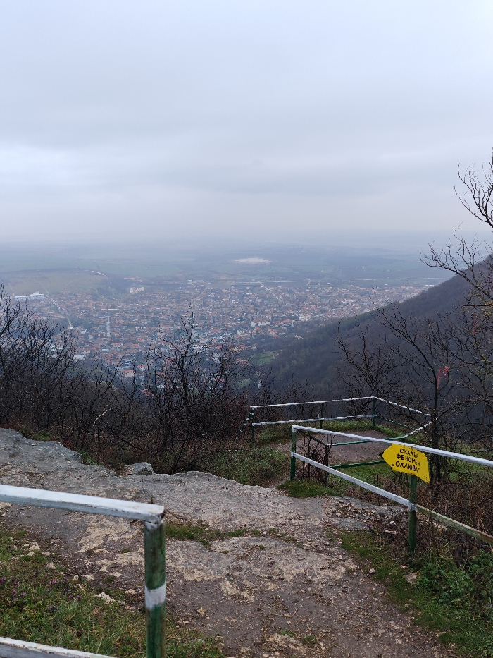 The scene depicts an elevated, panoramic view of the city of Shumen, Bulgaria, observed from a rocky and vegetated vantage point on an overcast day. In the immediate foreground, a rough, uneven ground surface of dark rock and soil, interspersed with sparse green and dried brown vegetation, is partially contained by a white and green metal railing system. The railing comprises white horizontal bars connected to posts painted green with white stripes. A yellow rectangular directional sign, featuring a right-pointing arrow and Cyrillic text "СКАЛЕН ФЕНОМЕН „ОХЛЮВЪ“" (Rock Phenomenon "The Snail"), is affixed to a railing post on the right side. Beyond the railing, a steep, dark green hillside, dotted with numerous leafless deciduous trees and bushes, descends into the urban area. The city of Shumen occupies the midground, presenting as a dense expanse of buildings, predominantly with reddish-brown roofs, along with visible street networks. In the distant background, the urban development transitions into flatter terrain with scattered structures and agricultural fields, leading to a large, light-colored, exposed earth area, consistent with a quarry or industrial site, on the far right. The upper third of the image is dominated by a uniformly gray, cloud-filled sky. No individuals are present, and no specific actions or interactions are visible within the frame.
