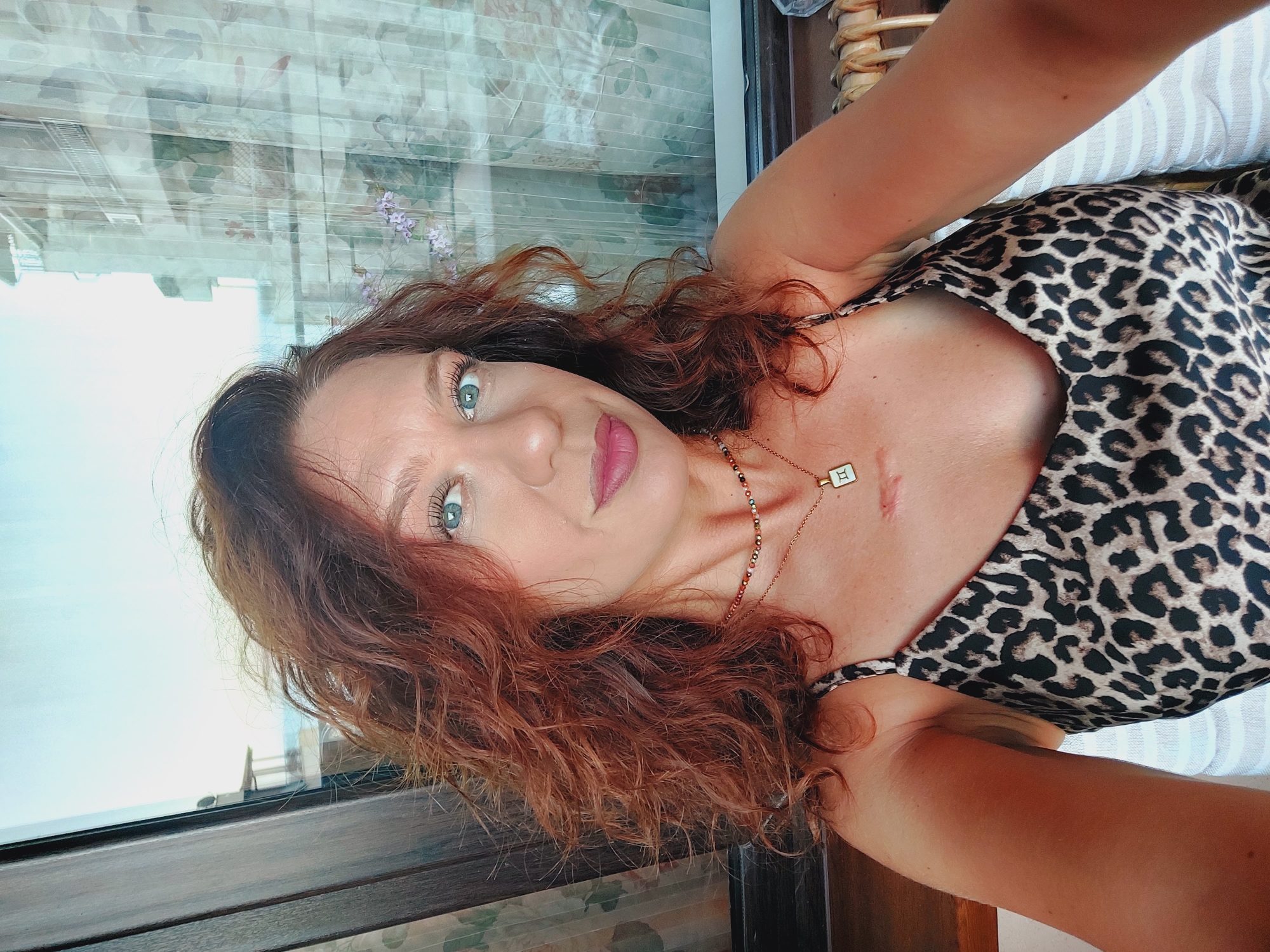A woman with red curly hair and blue eyes is taking a selfie in Ravda, Bulgaria. She is wearing a leopard print dress and a necklace with a square pendant. The background shows a window with curtains and a blurry view of buildings and the sea. She has a scar on her chest.