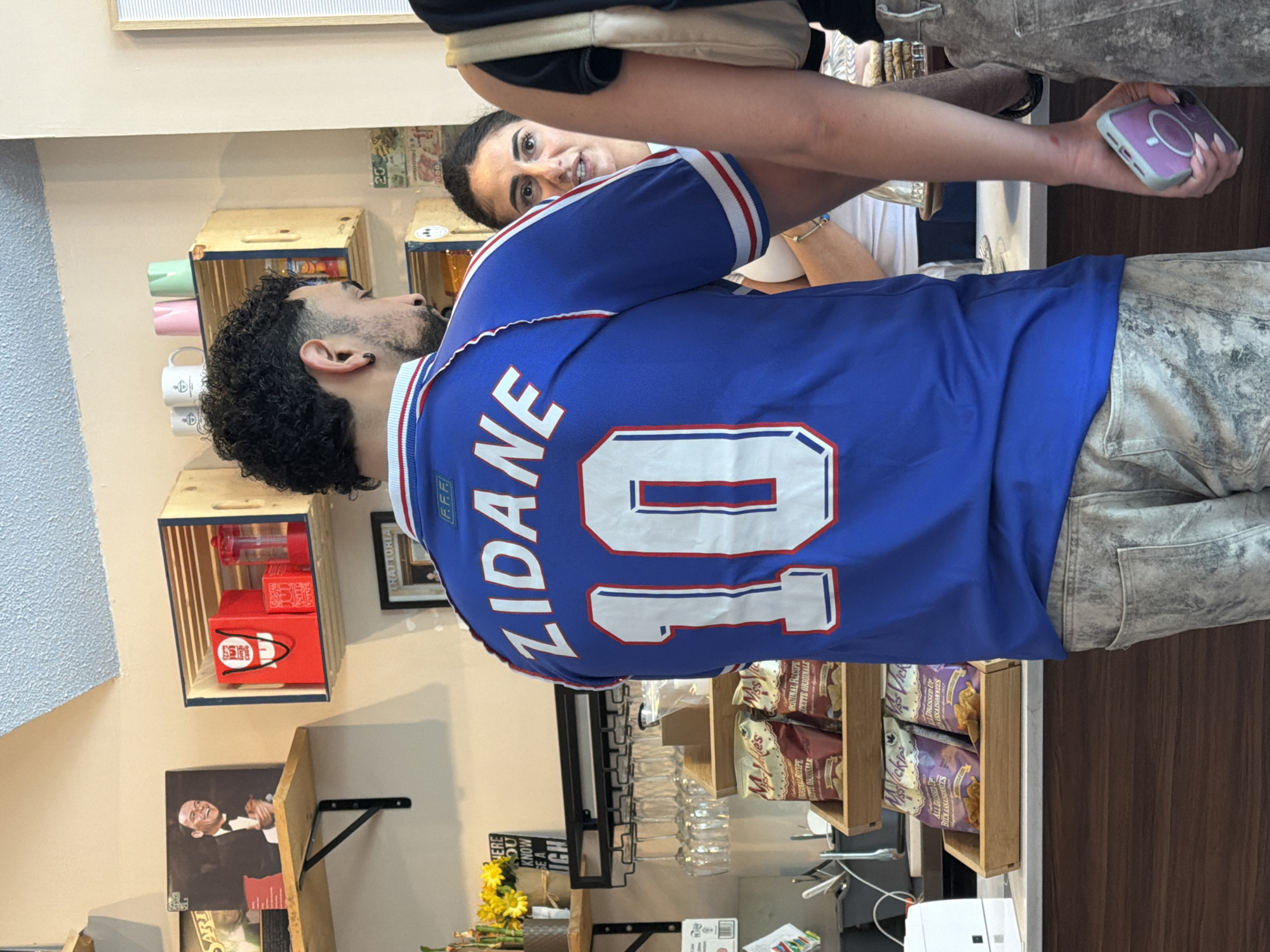 The image shows a medium shot of a man wearing a blue France national team jersey with "ZIDANE" and the number "10" on the back. He has curly hair and an earring in his left ear. A woman is partially visible in the background, looking towards the camera. Another person, partially obscured, is holding a smartphone.
The setting appears to be a cafe or deli. Wooden shelves and crates are mounted on the wall, displaying items like mugs, containers, and packaged food. There is a black-and-white portrait on the wall, and a bouquet of yellow flowers is on a counter. A wine glass rack with several glasses is also visible. Text on the wall includes "TRATTORIA", "KNOW", and "HERE YOU KNOW." Some text on the food packaging is "BIEN ASSAISONEES", "ORIGINAL RECIPE", and "ALL DRESSED UP". There is Canadian currency affixed to the wall.