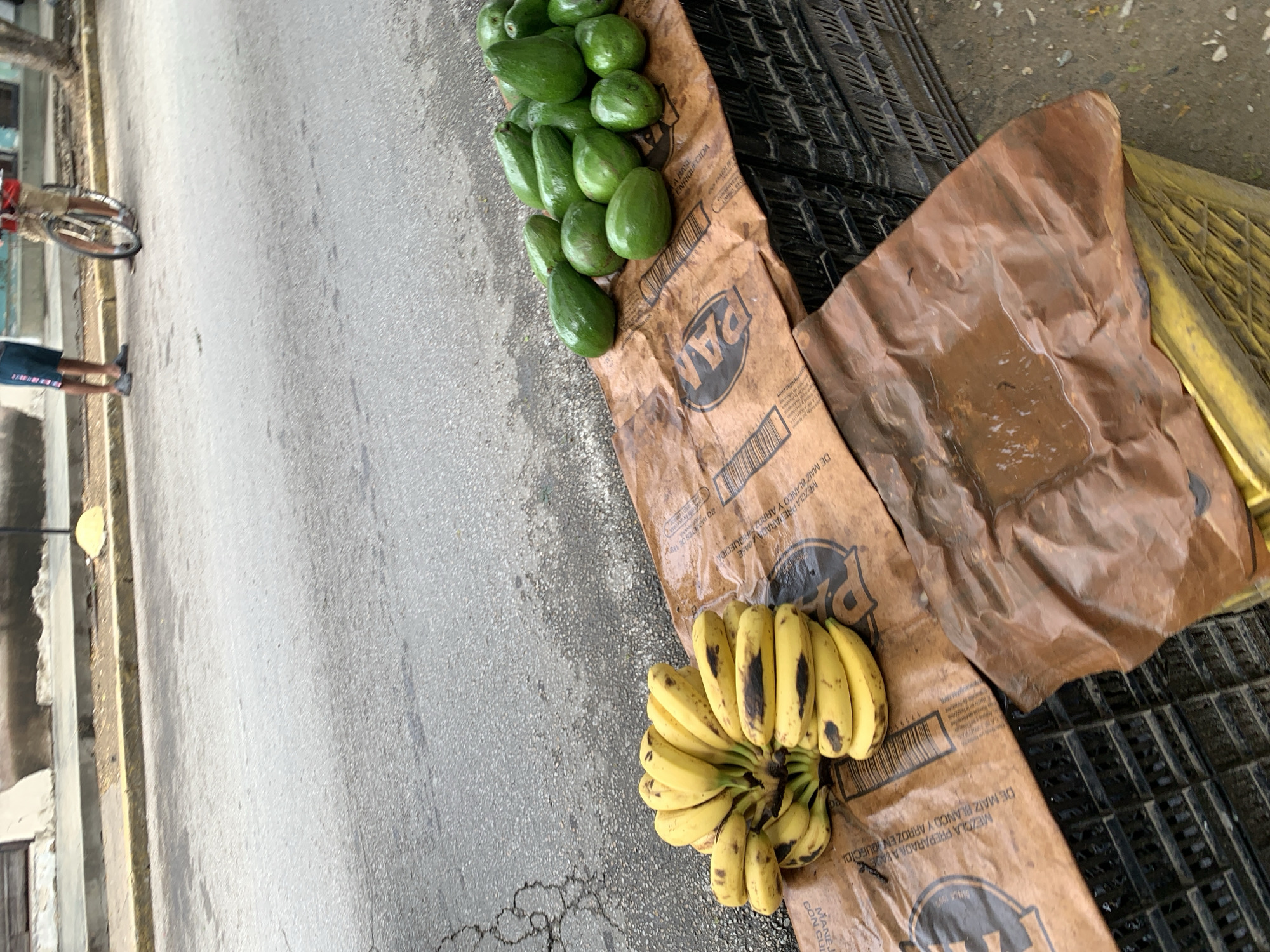 This image captures a vibrant roadside display of fresh produce in Santa Ana, Venezuela, likely during daylight hours. A collection of bright green avocados is neatly arranged on a brown paper bag, positioned on the edge of a paved street or road. Adjacent to the avocados, a large bunch of ripe yellow bananas, some with dark spots indicating ripeness, rests on another similar brown paper bag. These bags bear visible text, including the brand "PAN" and phrases such as "MEZCLA PREPARADA A BASE DE MAIZ BLANCO Y ARROZ ENRIQUECIDA" (prepared mix based on enriched white corn and rice), suggesting they are repurposed flour bags.
In the background, on the left side, the lower half of a person's legs, clad in shorts, can be seen next to a bicycle, indicating a pedestrian and cycling presence on the street. The road surface itself is asphalt, showing some cracks and light wear. To the far right, parts of black and yellow plastic crates are visible, possibly used for holding or transporting the produce. The scene depicts an informal street vending setup, a common sight for selling local produce in many regions. The clear lighting suggests fair weather, and the overall impression is one of a casual, everyday commercial activity.