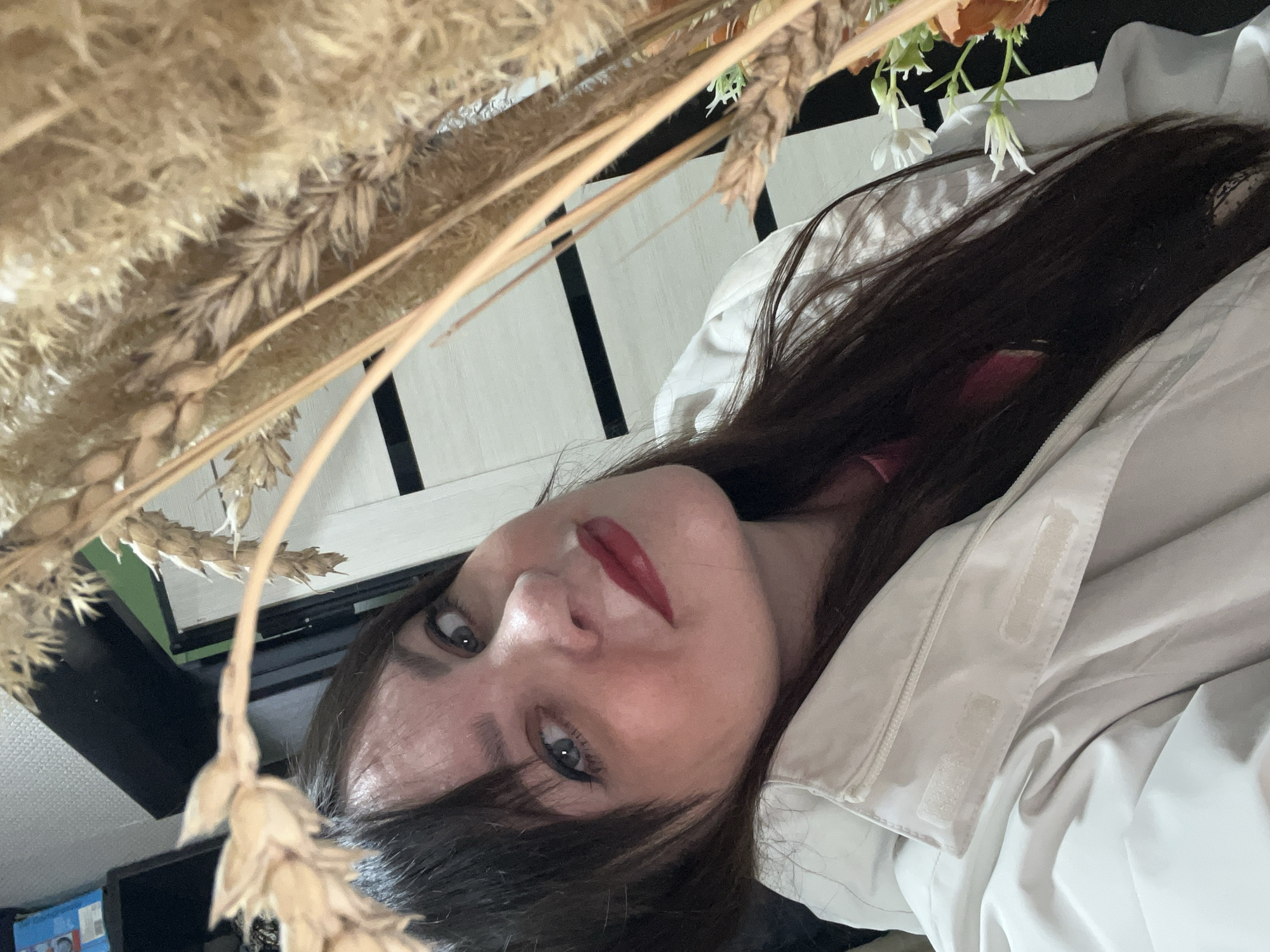 A woman with long, dark hair, red lipstick, and light-colored eyes looks at the camera. She is wearing a light-colored jacket or coat. The image is taken from a low angle, looking up at the woman. There are dried wheat stalks or similar grasses in the foreground, partially obscuring the view. Behind her, out of focus, is what appears to be a white wall with black vertical stripes. There is also a glimpse of a flower arrangement with white and orange flowers. Based on the information that this scene took place in the city of Noviy, Russia, it is reasonable to assume this is an indoor shot, possibly a home or office, with the woman posing with the wheat stalks or flowers as a prop.