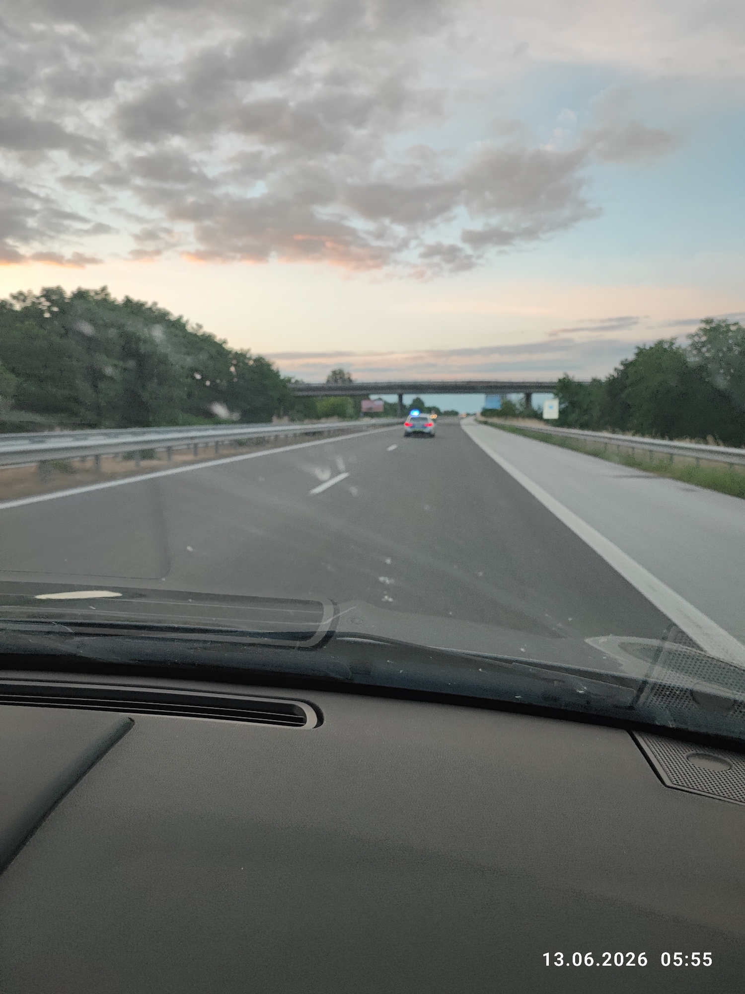 An image taken from inside a vehicle shows a highway at dusk. The sky is filled with clouds tinged with pink and orange from the setting sun. On the right side of the highway, a dense line of trees forms a barrier. In the distance, a highway overpass spans across the road, and a police car with flashing blue lights is visible on the highway ahead. The foreground of the image is dominated by the interior of the vehicle, including the dashboard and the windshield. The date and time stamp on the image reads "13.06.2026 05:55".