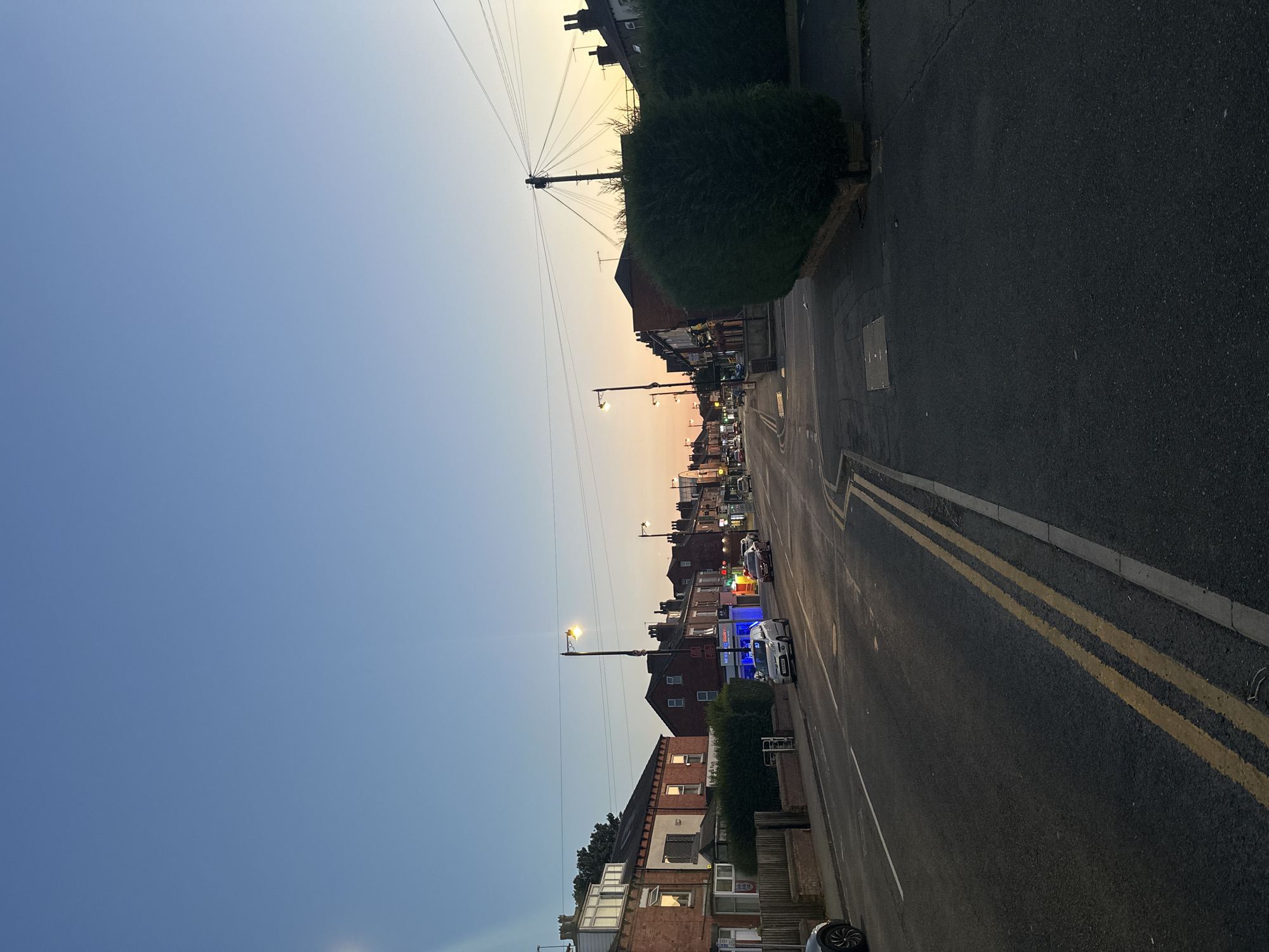 This is a medium shot, eye-level, outdoor photograph of a residential street in the city of Leeds, United Kingdom. The image depicts a view down a street lined with terraced houses and shops. The sky is a clear blue with a gradient to orange and yellow on the horizon, indicating either sunrise or sunset. The street is paved with asphalt and marked with double yellow lines in the foreground. There are several cars parked and driving on the street. On the left side of the street, there are brick-built houses with windows and chimneys, and a neatly trimmed hedge. On the right side of the street, there are more shops with illuminated signs, a large pruned hedge, and a lamppost. Power lines are visible stretching across the scene, supported by poles. The overall atmosphere is tranquil and captures a typical urban street scene at dusk or dawn.