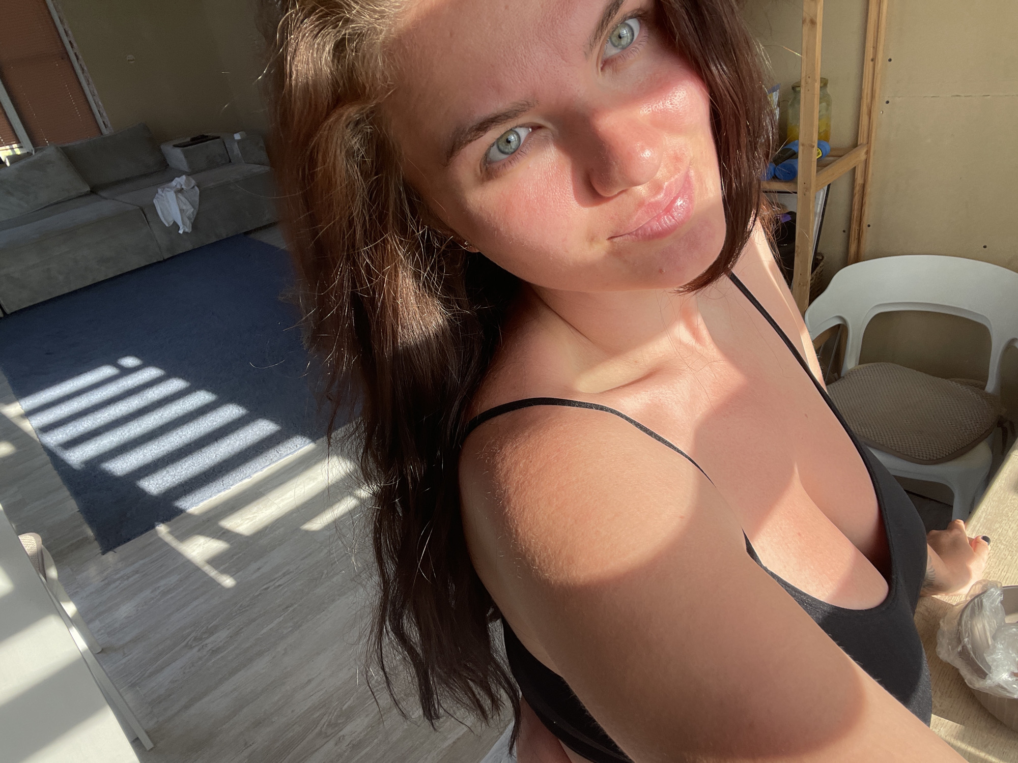 A selfie captures a woman with blue eyes and brown hair, wearing a black tank top. The lighting is bright, with shadows falling across her face and body. Behind her, a room is visible, including a sofa, a rug, and a white chair near a table. The location is suggested as Мосметаллоконструкция Онт, Russia.