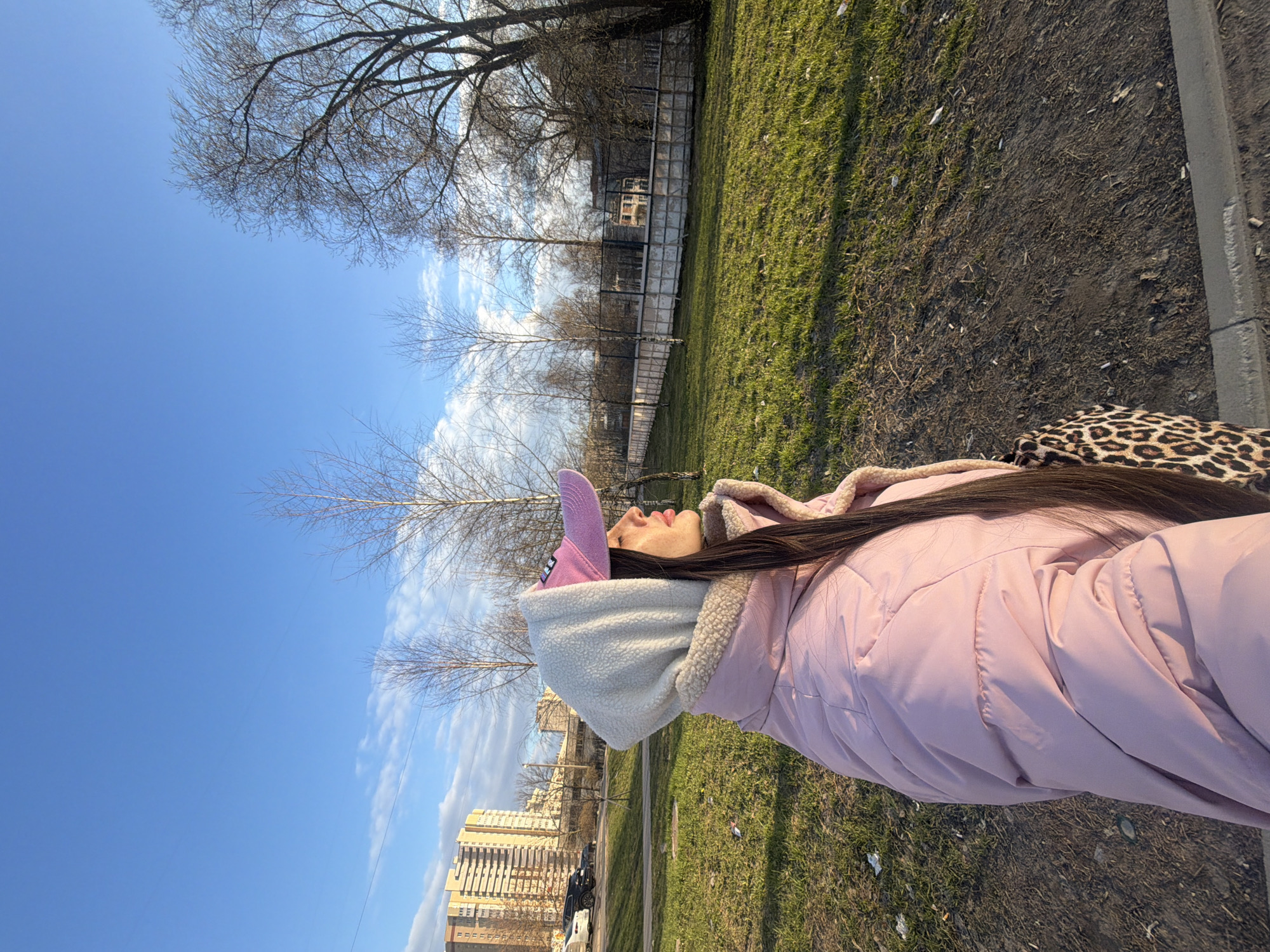 A woman wearing a pink jacket with a furry-lined hood and a pink cap with a pointed horn is standing on a grassy area next to a paved path. She is looking up at the sky. A building can be seen in the background. The image was taken in Saint Petersburg, Russia.