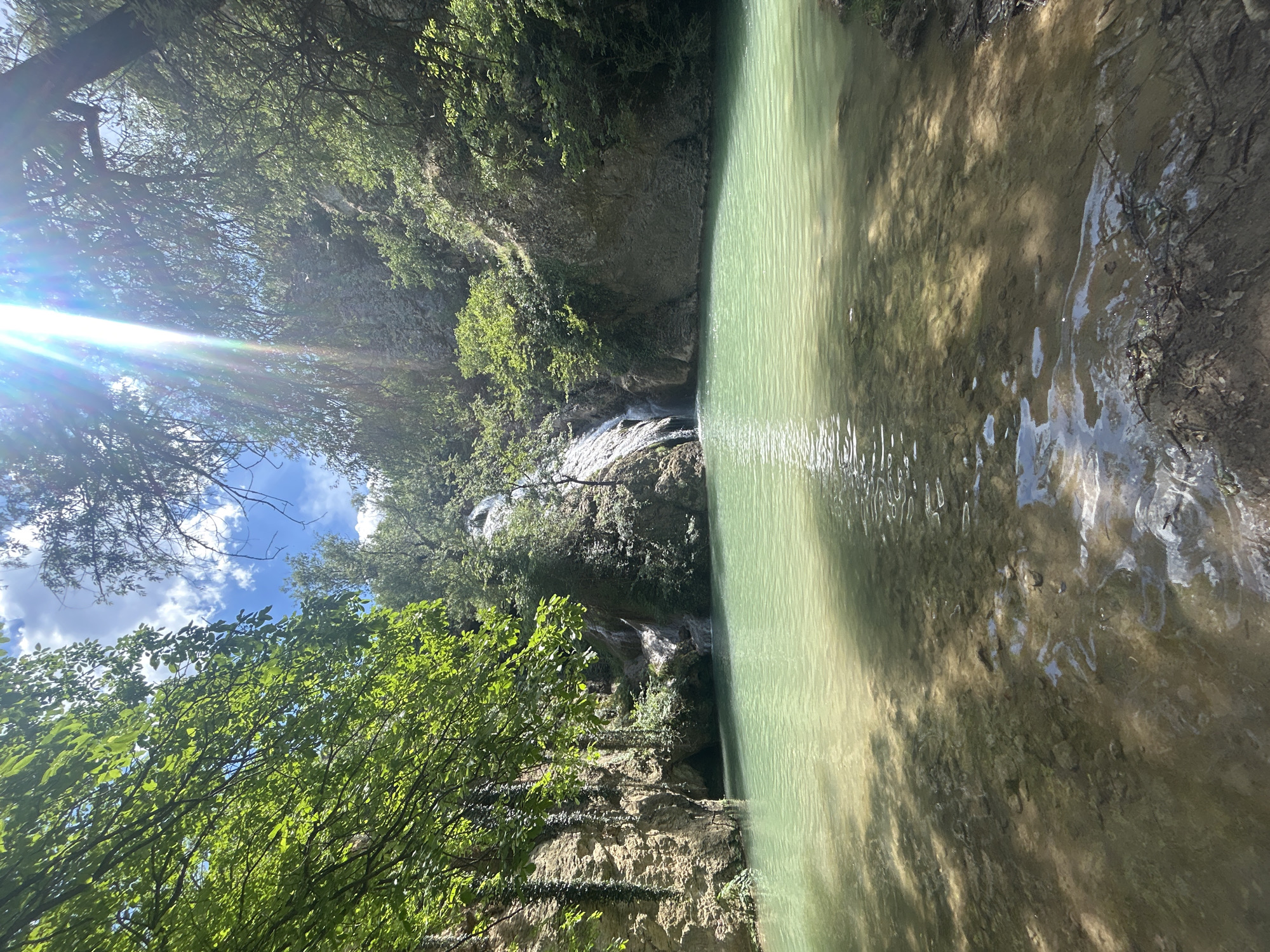 The image depicts the "Krushuna Falls," a picturesque waterfall located in the village of Hotnitsa, Bulgaria. The scene is dominated by a wide pool of clear, turquoise water in the foreground, with a waterfall cascading down a rocky cliff face in the background. Lush green vegetation covers the surrounding cliffs and trees, with sunlight filtering through the leaves, creating dappled shadows on the water. The water is shallow near the shore, revealing the rocky bottom, and deepens towards the pool. The overall impression is one of natural beauty and tranquility.