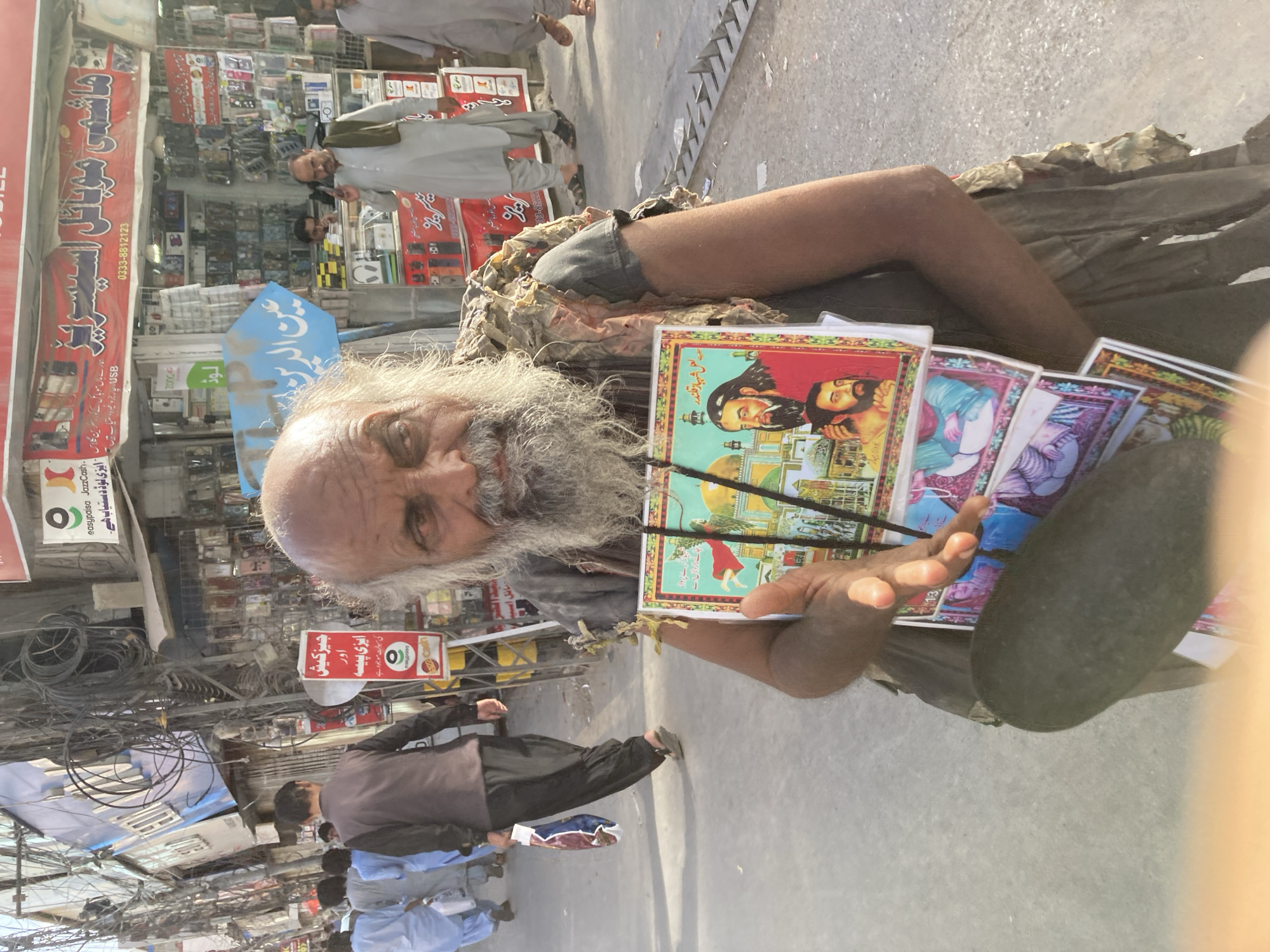 The image depicts a man with a long white beard sitting on the ground in what appears to be a busy street in Quetta City Tehsil, Pakistan. He is holding up a stack of cards or pictures, one of which features an image of two men, possibly historical figures. In the background, there is a shop with signage in Urdu, indicating mobile phone services. Overhead, a dense network of cables and wires is visible. People are walking by in the street, and other shops are visible in the background.