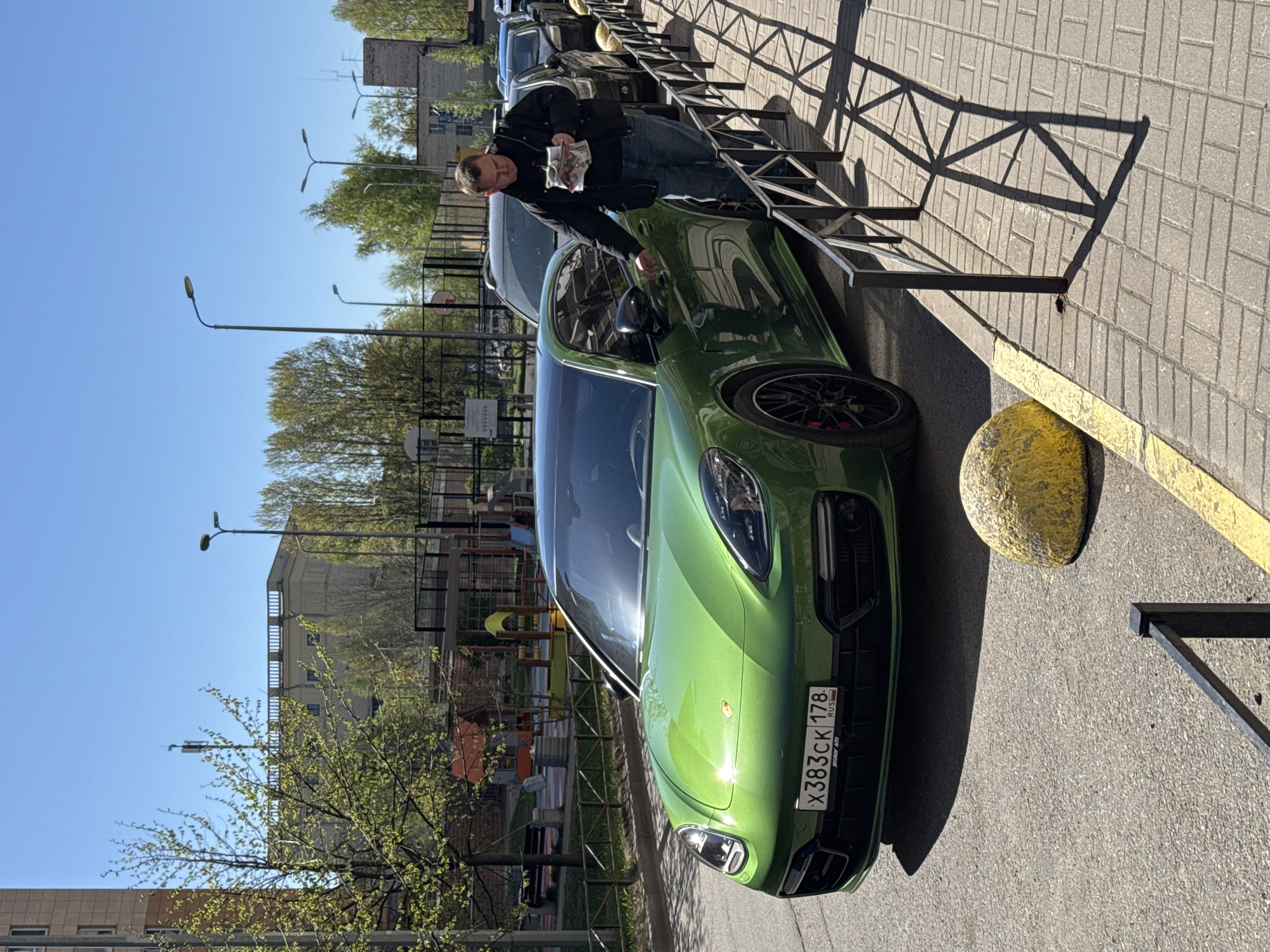 A man wearing a black jacket and jeans is standing next to a green Porsche Panamera with the license plate "X383CK 178 RUS." He is holding what appears to be Russian currency in his hands and appears to be reaching into the car. The car is parked on a gray brick sidewalk next to a black metal fence and a round yellow concrete barrier. In the background, there are trees, buildings, streetlights, and a playground. The photo appears to be taken during the daytime in Vsevolozhsk, Russia.