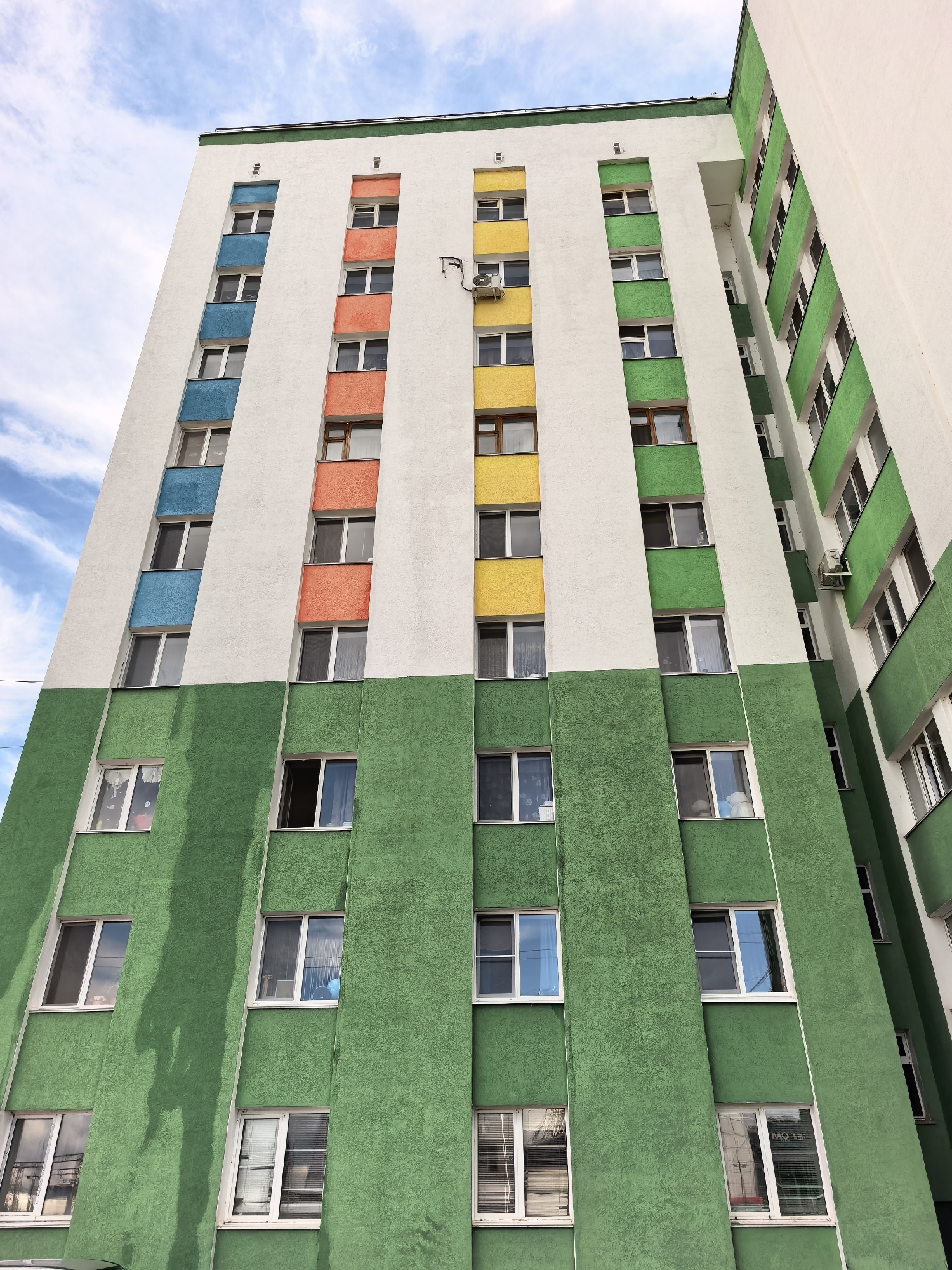 A tall, multi-story residential building in Sterlitamak, Russia, is captured from a low-angle perspective, emphasizing its height against a partly cloudy blue sky. The building's facade features a striking color scheme. The lower section, comprising several floors, is a textured, vibrant green, with vertical green strips framing windows. Above this, the facade transitions to a clean white, which is visually segmented by vertical bands of distinct colors: blue, orange, yellow, and green. Each band contains multiple windows, many reflecting the bright sky. An outdoor air conditioning unit is mounted on the white section of the facade.
The setting is clearly daytime, with ample sunlight illuminating the building and a sky exhibiting soft white clouds against a broad blue expanse. No human activity is directly visible, but the numerous windows suggest residential occupancy. The green portion of the building shows some streaking, possibly from water runoff. Reflections in some lower windows on the green facade reveal partial, mirrored text, including "MO" and "FELOW." The overall impression is of a modern or renovated apartment block, adding a colorful element to the urban landscape.