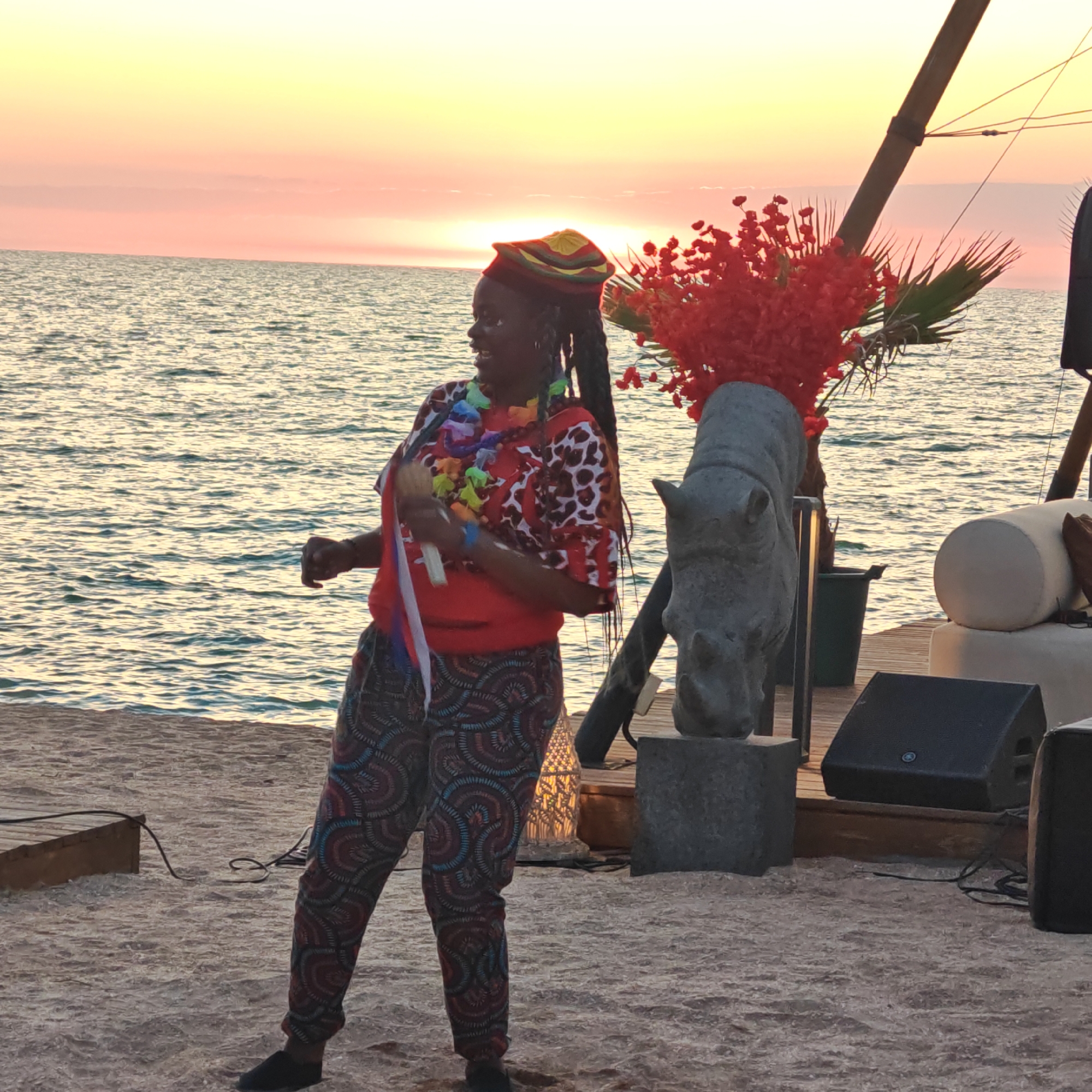 The image shows a woman standing on a sandy beach at sunset. She is wearing a colorful hat, a red shirt with a leopard print pattern, and patterned pants. She is holding a microphone and appears to be singing or performing. In the background, there is the ocean and a sunset with vibrant orange and yellow hues. To the right of the woman, there is a stone sculpture of a rhinoceros head and some red flowers. There are also speakers and other equipment on a wooden platform, suggesting an outdoor event or performance. The location is identified as Мысовское сельское поселение, Ukraine.