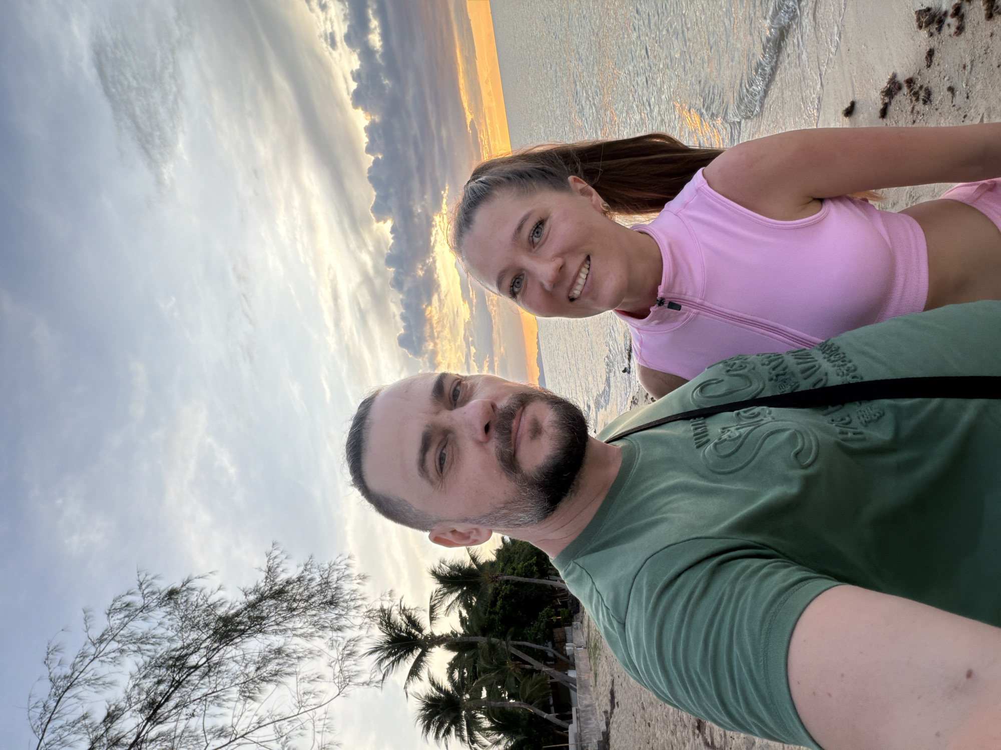 Two individuals, a man and a woman, are captured in a selfie on a beach at sunset in Baan Thong Po, Thailand. The man, on the left, has short dark hair and a beard, and is wearing a green t-shirt. The woman, on the right, has long brown hair in a ponytail and is wearing a pink sleeveless top. They are standing on a sandy beach with the ocean and a sunset sky in the background. Palm trees are visible behind them to the left. The tide is low, with gentle waves lapping at the shore. The overall scene suggests a relaxed and pleasant atmosphere during twilight.