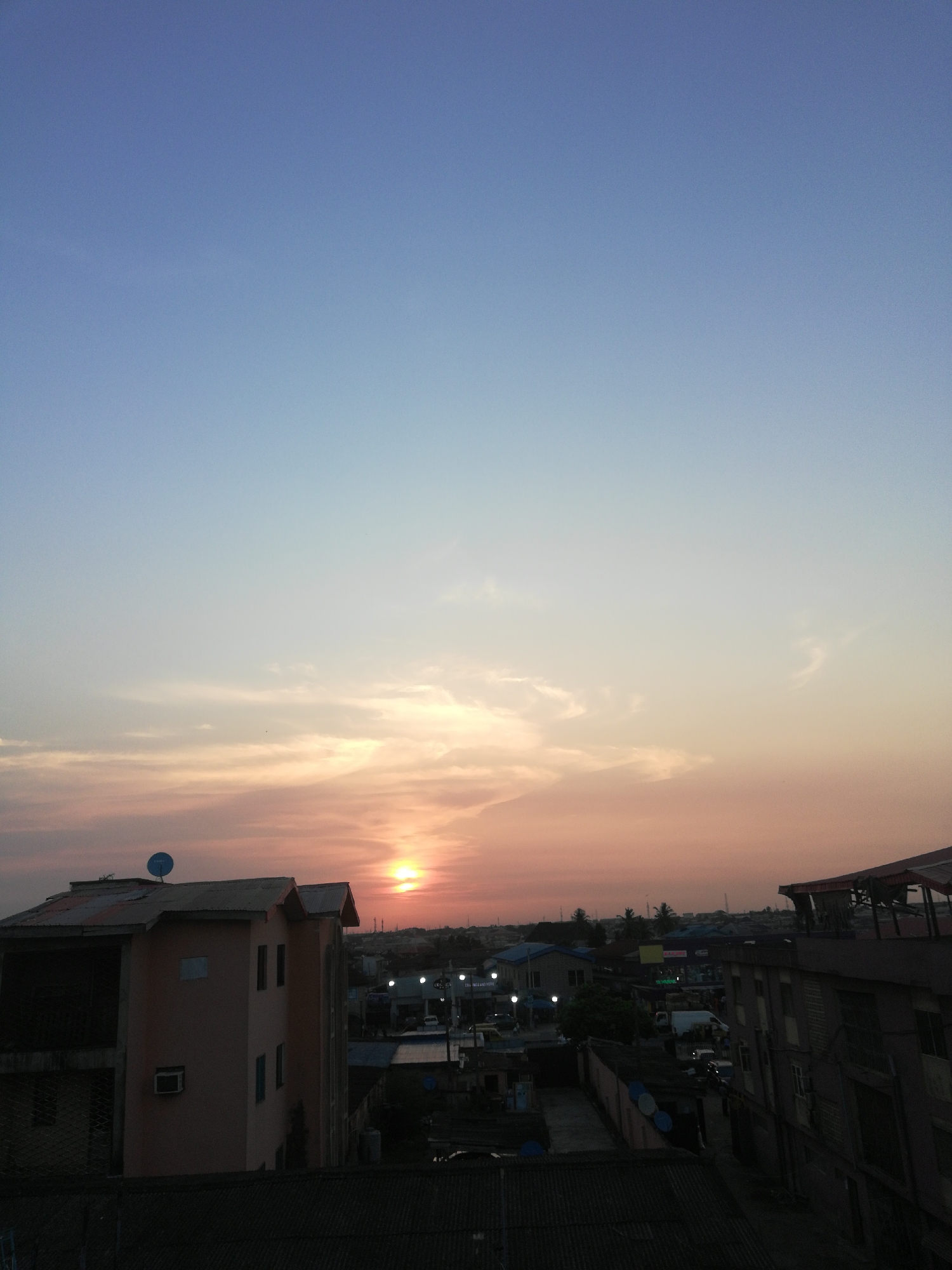 The image presents an expansive view of an urban landscape at sunset, consistent with a setting in Lagos, Nigeria. The sky occupies the majority of the frame, showcasing a dramatic gradient of colors, transitioning from deep blue at the zenith, through lighter blues and soft purples, to warm oranges and pinks near the horizon where the sun is setting. The sun itself is a vibrant orange orb, low in the sky and slightly to the left of the center, casting a warm glow and illuminating scattered, wispy clouds.
In the foreground, the dark silhouettes of building roofs create a stark contrast against the bright sky. On the left, a prominent multi-story building, appearing light peach or beige, features a distinctive corrugated roof and a blue satellite dish. An air conditioning unit is also visible on its facade. On the right, part of another building with a dark, angled roof is visible. The mid-ground reveals a dense urban environment characterized by numerous residential or commercial structures, many with corrugated iron roofs, indicative of a bustling city. As dusk settles, artificial lights from street lamps or vehicles begin to punctuate the urban tapestry. No specific human activity is discernible, and no individuals are clearly visible. The overall mood is one of quiet transition from day to night, with the natural beauty of the sunset contrasting with the man-made environment. No visible text is present in the image.