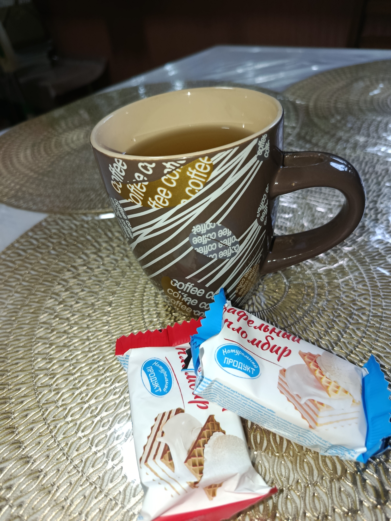 The image shows a close-up of a brown mug filled with a light-colored liquid, possibly tea or coffee, with text that reads "coffee" repeated around it. Two individually wrapped wafer snacks with cream filling and chocolate drizzle are placed in front of the mug on a golden, textured placemat. The snacks have blue and white packaging with Russian text, including "Натуральный ПРОДУКТ" (Natural PRODUCT) and "Сливочный пломбир" (Creamy ice cream). The background is blurred, but appears to be an indoor setting with a wooden surface. The overall scene suggests a moment of relaxation or a snack break. There is no visible indication of the location being Tashkent, Uzbekistan.