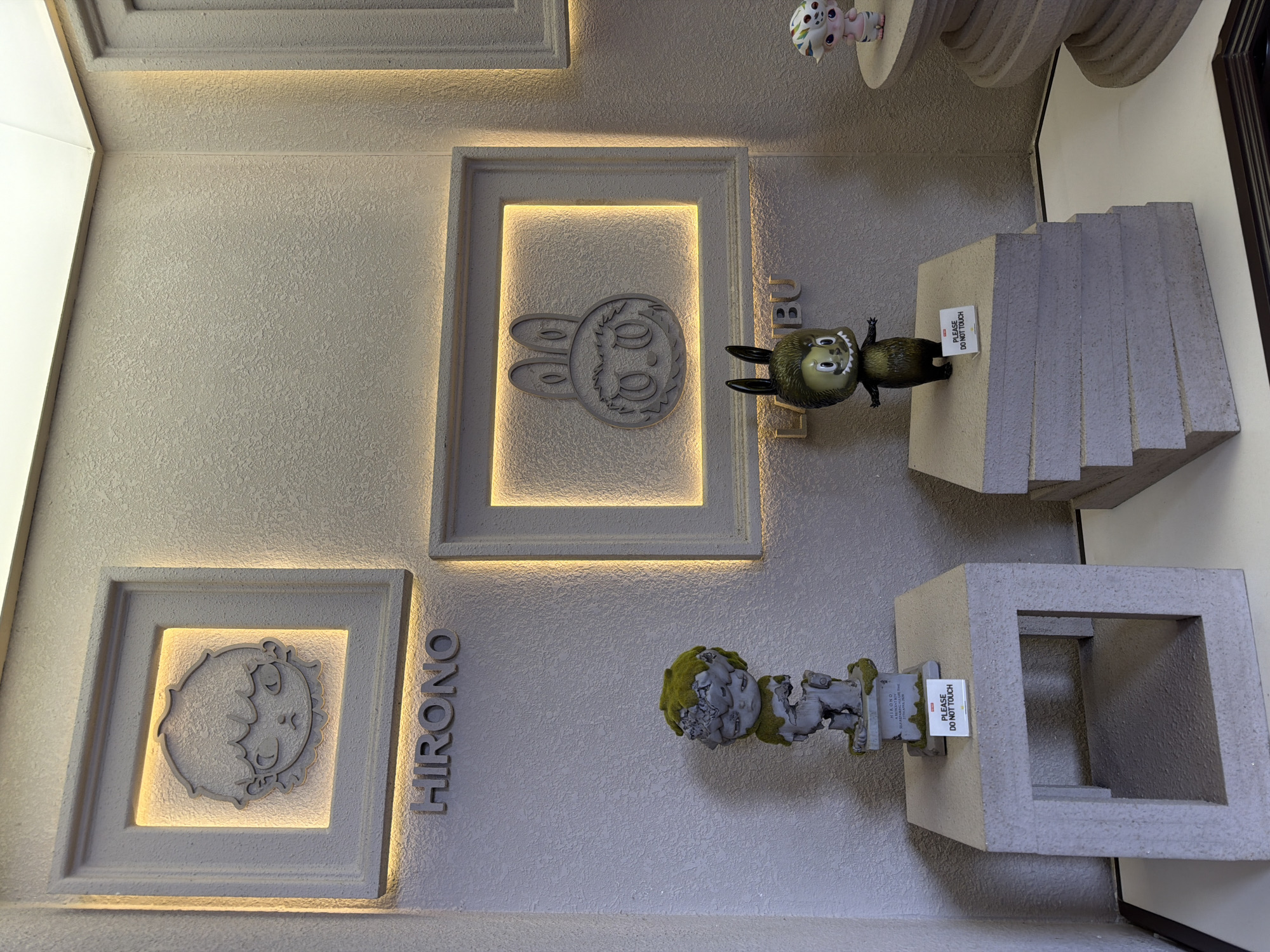The image shows a close-up of a wall display in what appears to be a shop or gallery. The wall is light gray with a textured surface. There are three framed artworks illuminated from behind. The one on the left displays a child's face, the one in the middle displays a rabbit, and the name "HIRONO" is written vertically next to the left frame.
To the right of these wall-mounted artworks, there are three-dimensional objects on stands. The top object is a small figurine of a green and black character, with a sign that reads "PLEASE DO NOT TOUCH". Next to it is a set of gray, block-like steps. Below this arrangement is a similar setup with another small figurine covered in green moss, also with a "PLEASE DO NOT TOUCH" sign, and a hollow gray cube. The image is likely taken in Đà Nẵng, Vietnam, based on text on one of the signs, although it is not explicitly stated.
