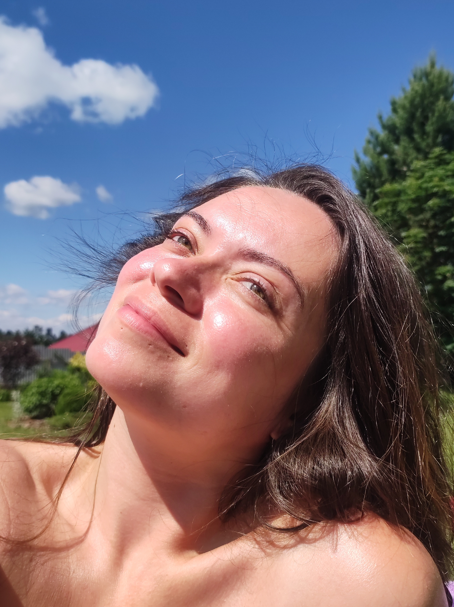 A close-up, low-angle, outdoor, waist-up shot features a young adult woman with brown hair and light skin, illuminated by bright sunlight. She is looking upwards, with her face tilted towards the sky. Her eyes are open, and she has a slight smile on her lips. Her skin appears flushed and shiny, likely due to the sun. Some strands of her hair are caught by the wind and are visible across her face.
In the background, a clear blue sky with scattered white clouds dominates the upper portion of the image. To the right, the upper part of a tall, green evergreen tree is visible. The lower background shows hints of a residential area, with greenery, bushes, and the peak of a red-tiled roof suggesting a house. The overall environment appears to be a garden or backyard. The image was likely taken in the city of Fokino, Russia, given the context provided.