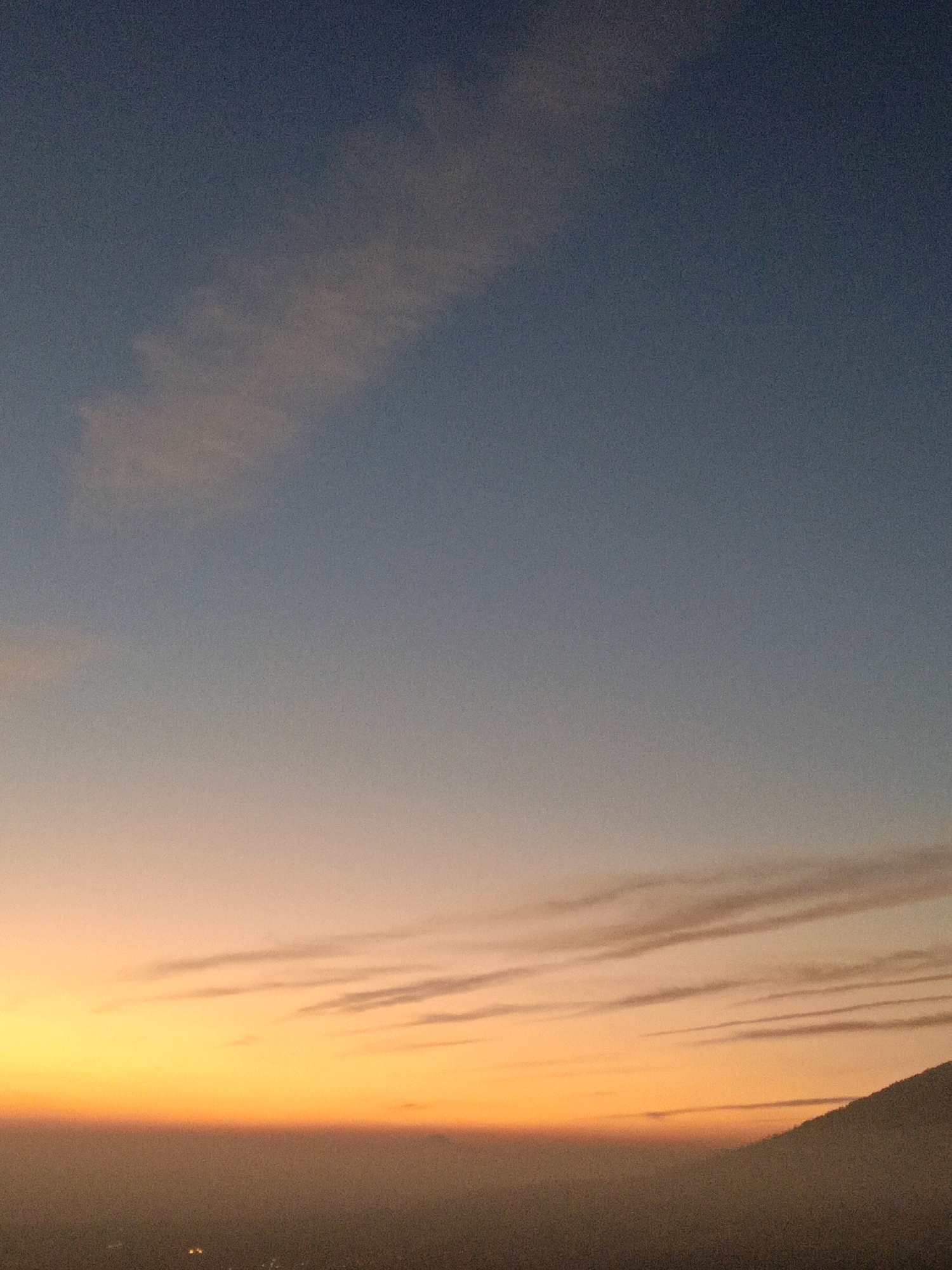 This is a vertical, full-frame, outdoor photograph taken at dusk. The image captures a gradient of colors in the sky, transitioning from a deep blue at the top to vibrant oranges and yellows near the horizon. Wispy clouds are scattered across the sky, with more prominent, elongated streaks of cloud present in the lower half of the image. In the lower right corner, the silhouette of a mountain or hill is visible, partially obscured by a hazy layer that extends across the lower portion of the frame. Beneath this haze, faint lights suggest the presence of a city or settlement. Specifically, this scene is located in Butuh Kidul, Indonesia.